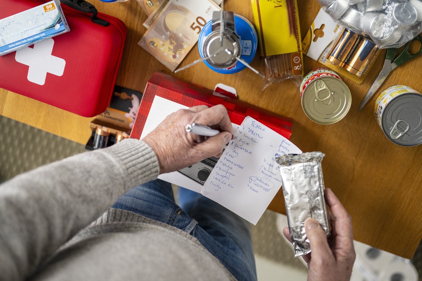 Person makes list of emergency supplies on wooden table with canned goods and red first aid kit. Preparing a survival weekend