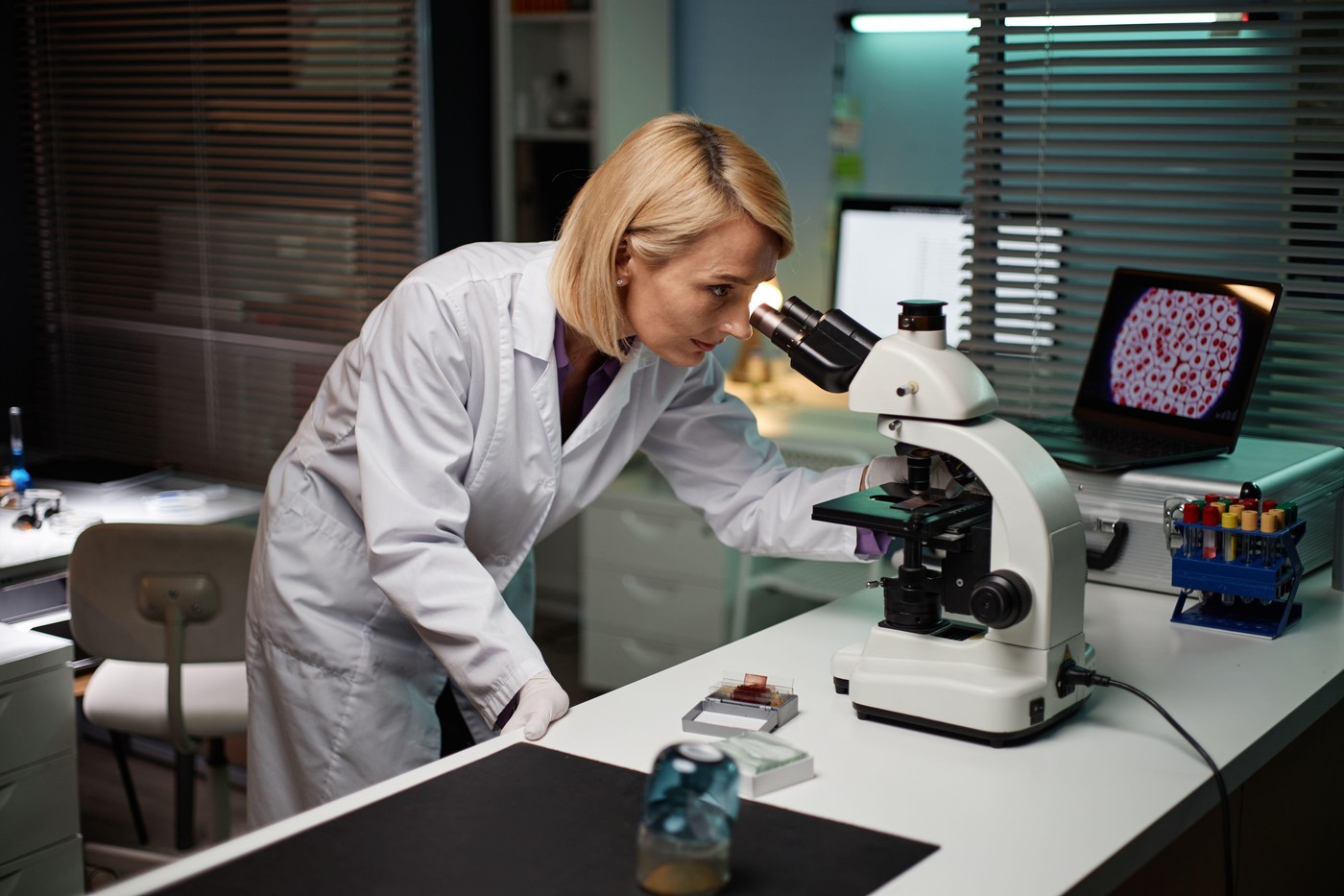 Middle aged Caucasian woman examining sample through microscope in scientific laboratory, focusing on research process with laboratory equipment and computer displaying cell images nearby