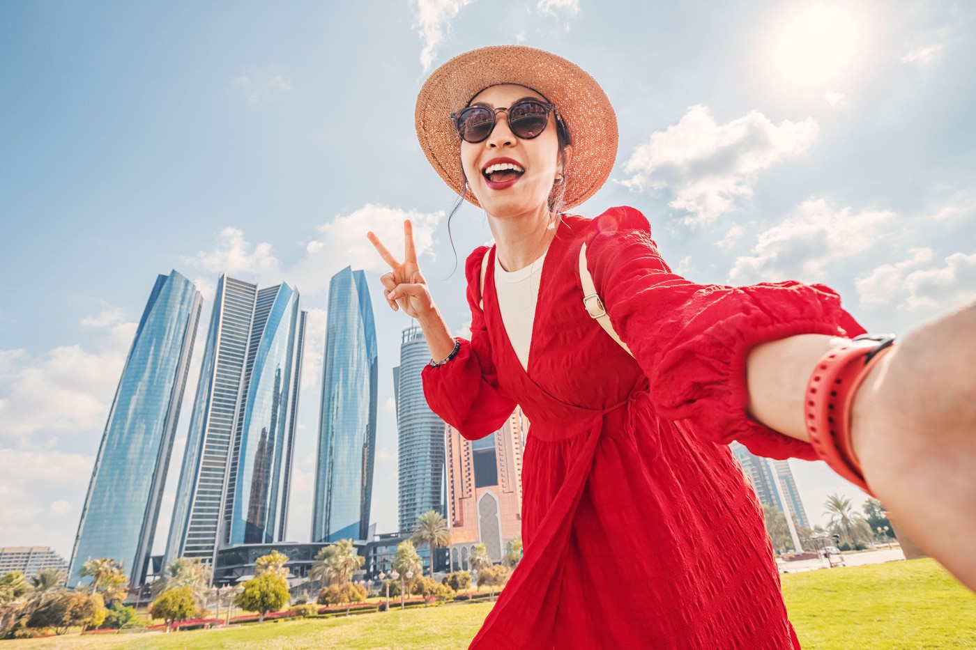 Young woman wearing red dress and straw hat taking a selfie with skyscrapers in the background in Abu Dhabi