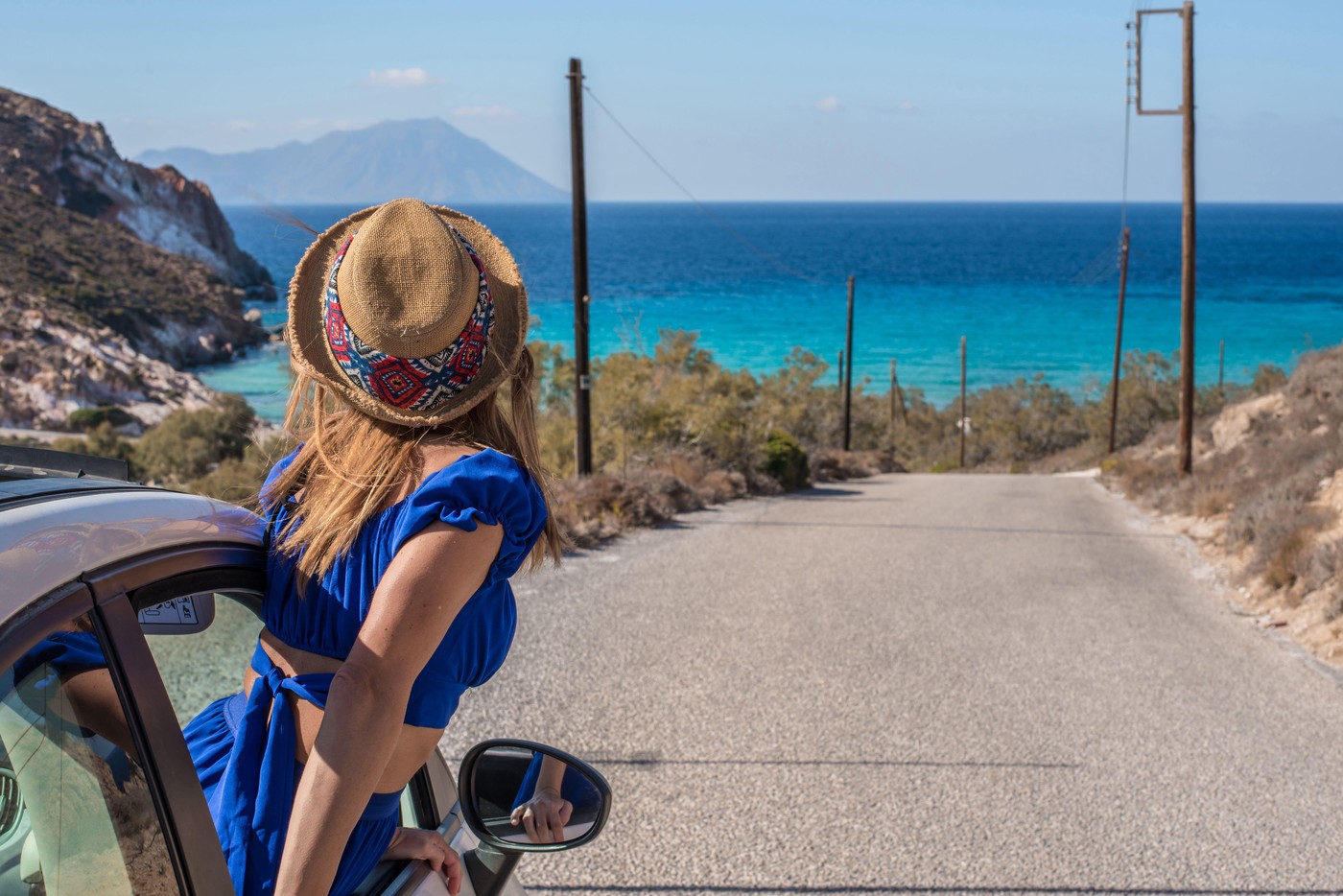 Woman in a blue dress and straw hat enjoys a scenic drive on Plathiena Road, Milos Island, Greece. The crystal-clear water and mountainous backdrop cr