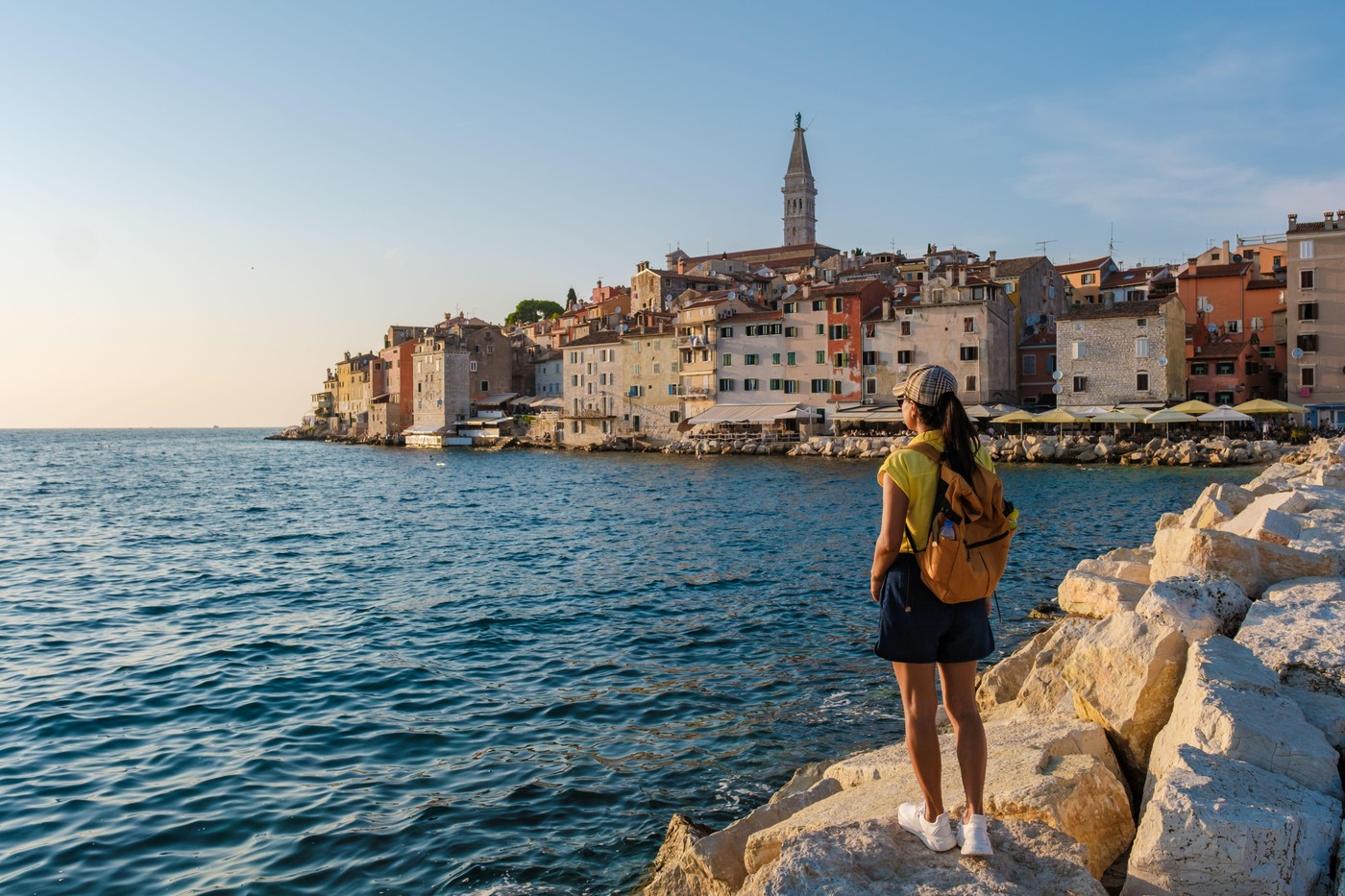 A traveler admires the stunning coastal view of Rovinj, Croatia, as golden sunlight illuminates the historic buildings and calm waters, creating a tranquil atmosphere at dusk.
