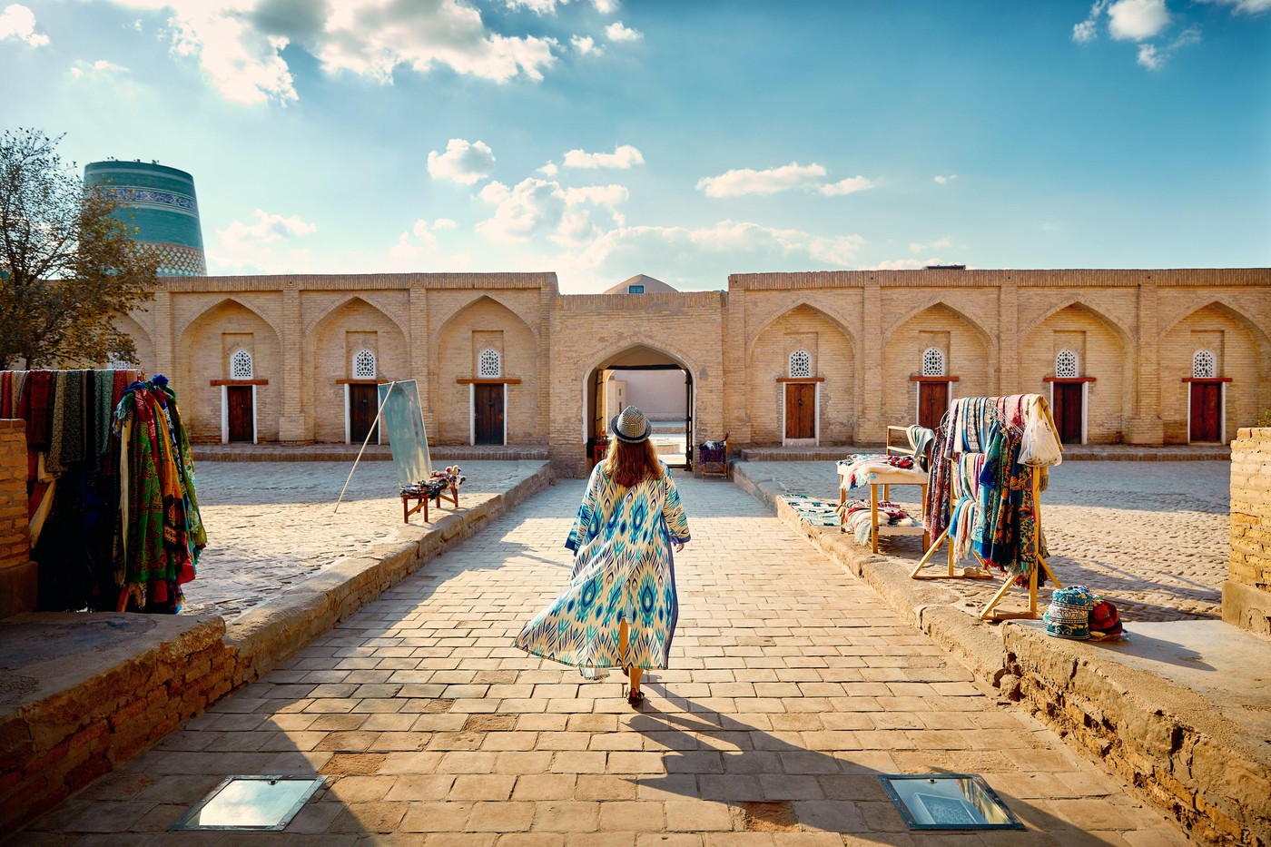 Young woman in ethnic dress with blue ornament and hat walking at street market near Kalta minor minaret at ancient city Itchan Kala of Khiva in Uzbek