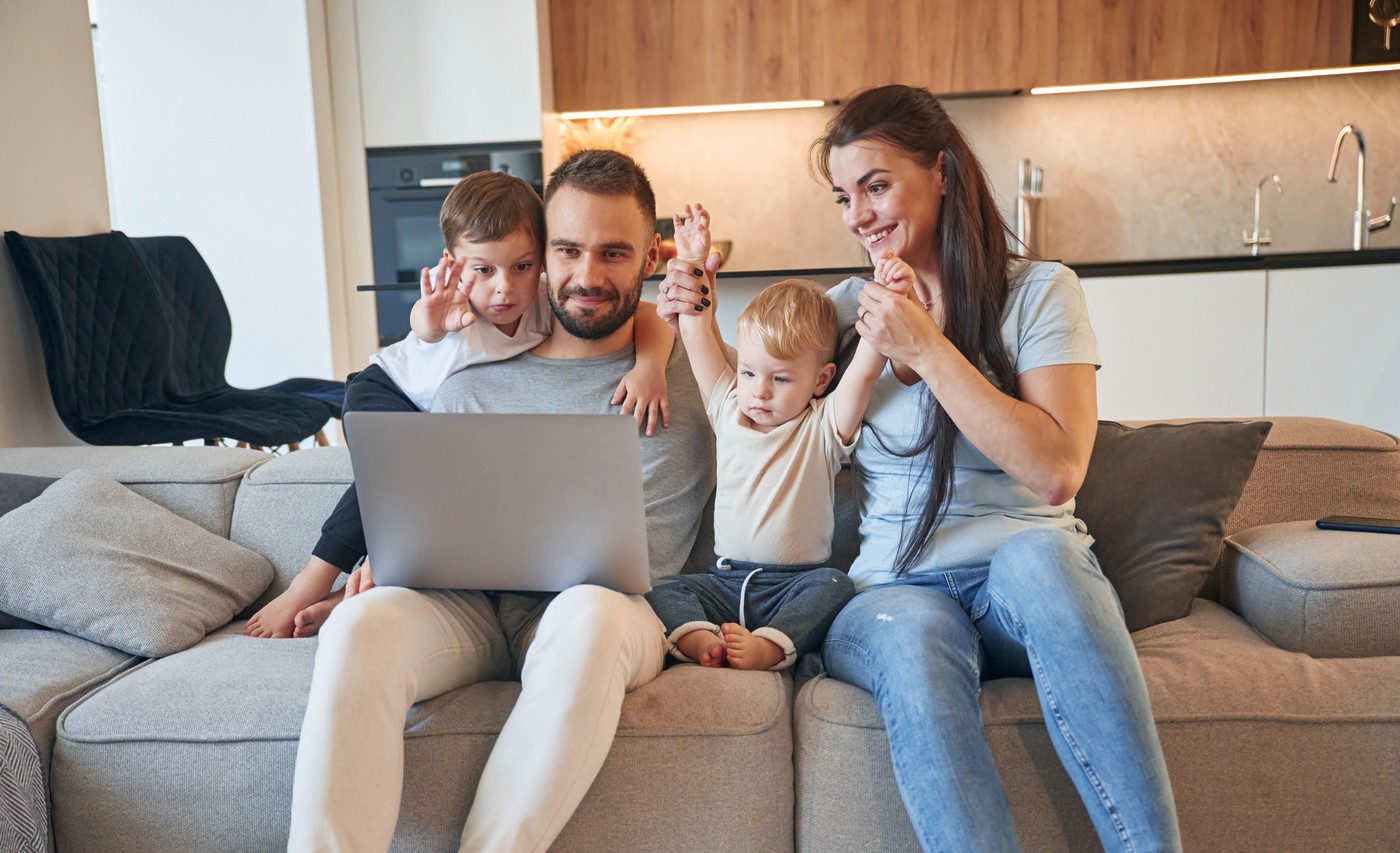 Silver colored laptop on legs. Parents with two little boys kids are together in domestic room.