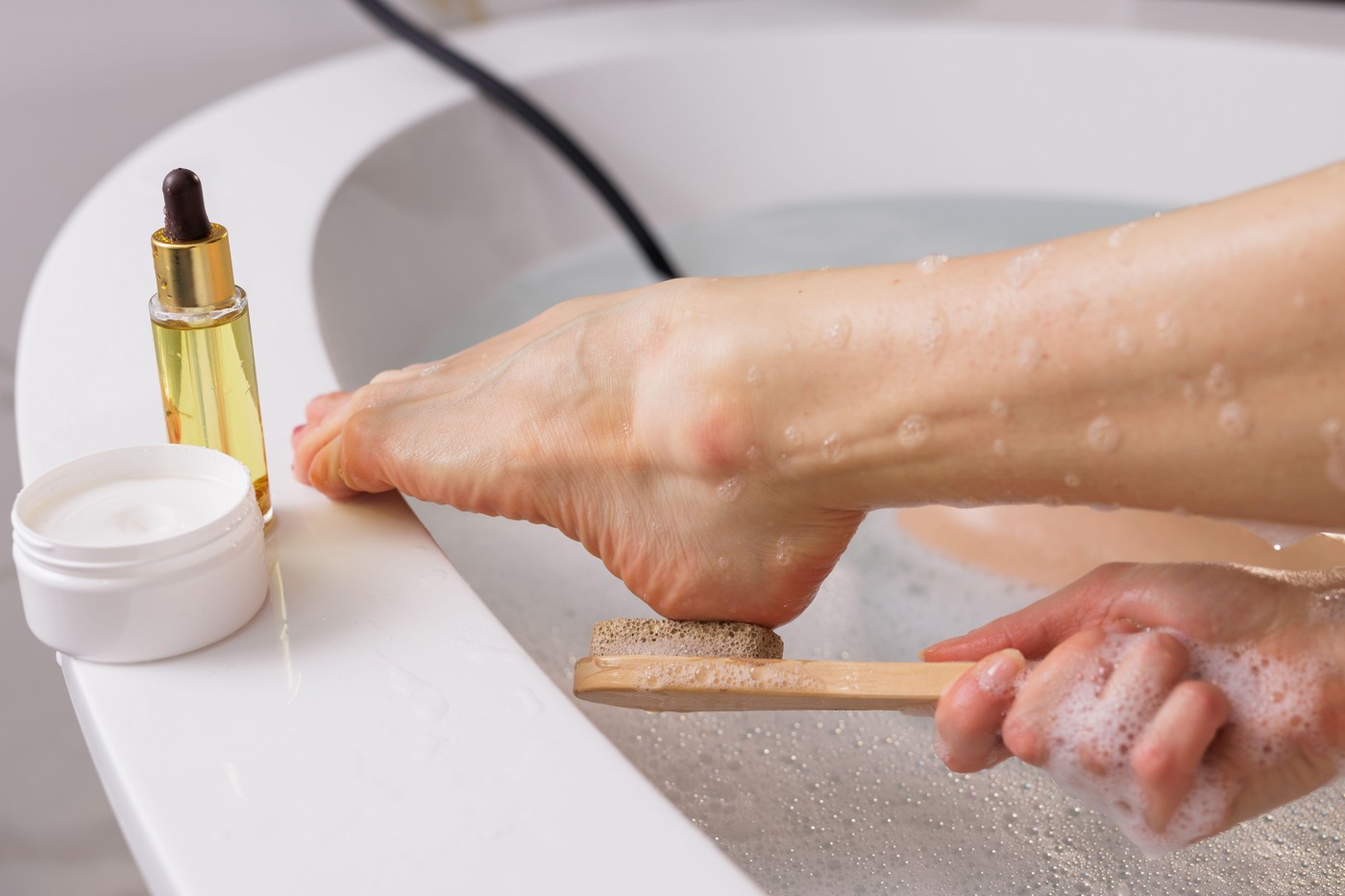 Foot care and relaxation. Woman exfoliating foot with a pumice stone in a warm bath. Skincare essentials like cream and oil nearby. Spa, self-care, an