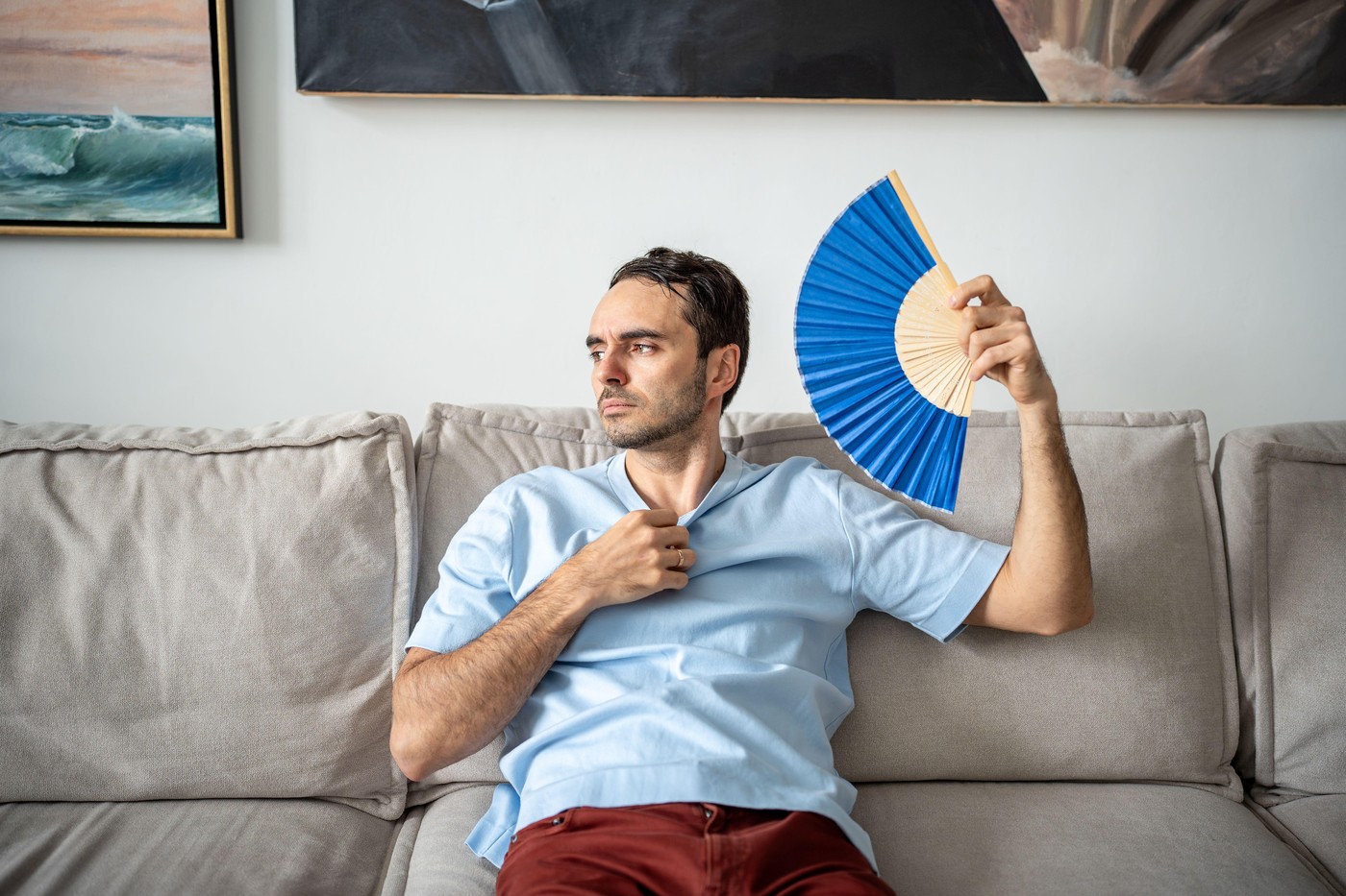 Sad man suffering from heat in apartment, fanning himself with paper fan, no air conditioner at home