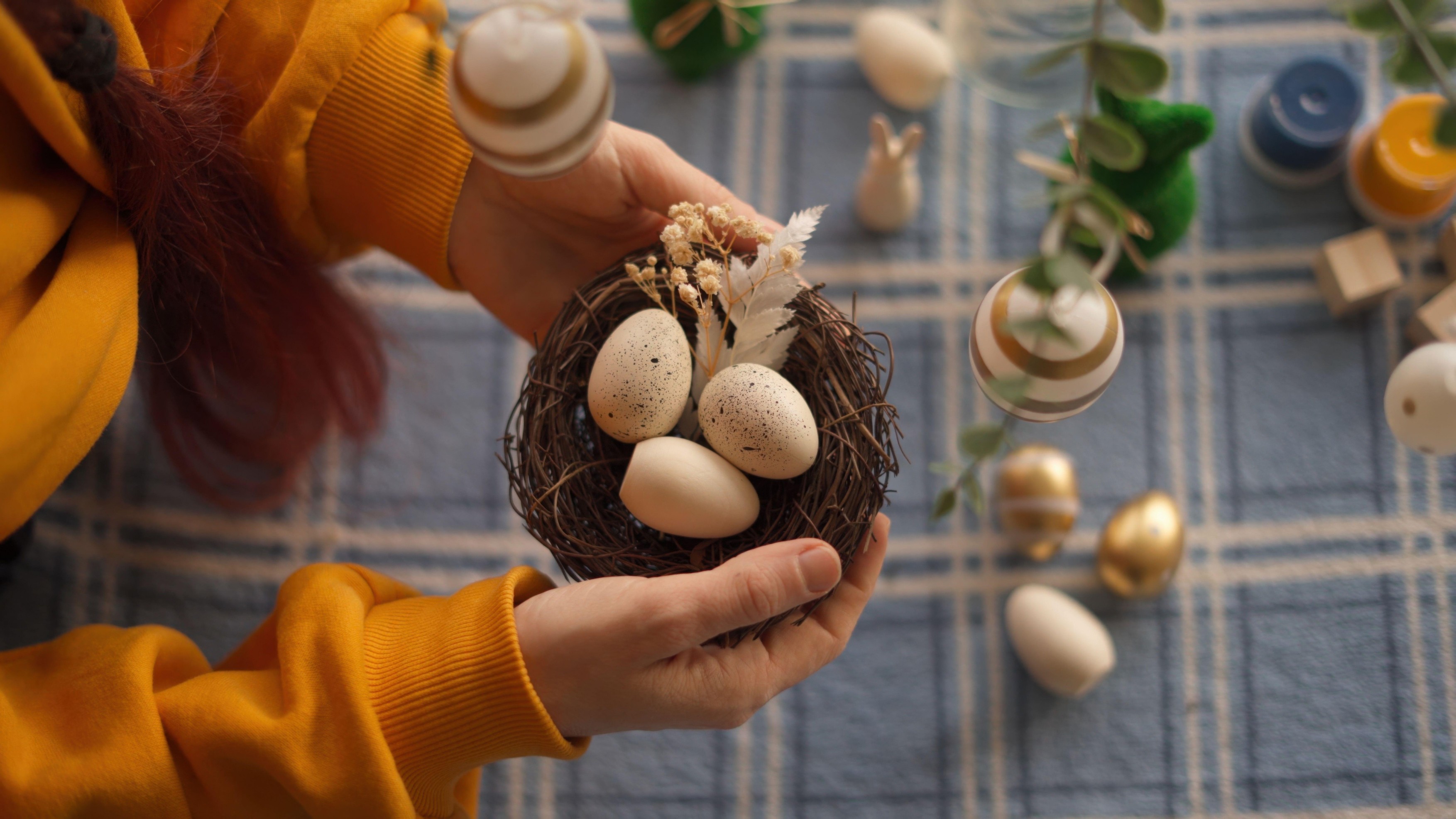 Person holding a nest with speckled eggs on a blue plaid tablecloth, surrounded by Easter decorations.,Image: 985322178, License: Royalty-free, Restrictions: , Model Release: no, Credit line: Oksana Hranovska / Alamy / Profimedia