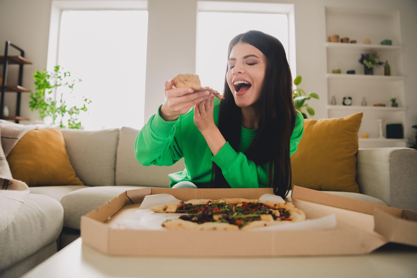 Young woman in a green pullover enjoying pizza indoors in a modern living room, showcasing a cheerful moment