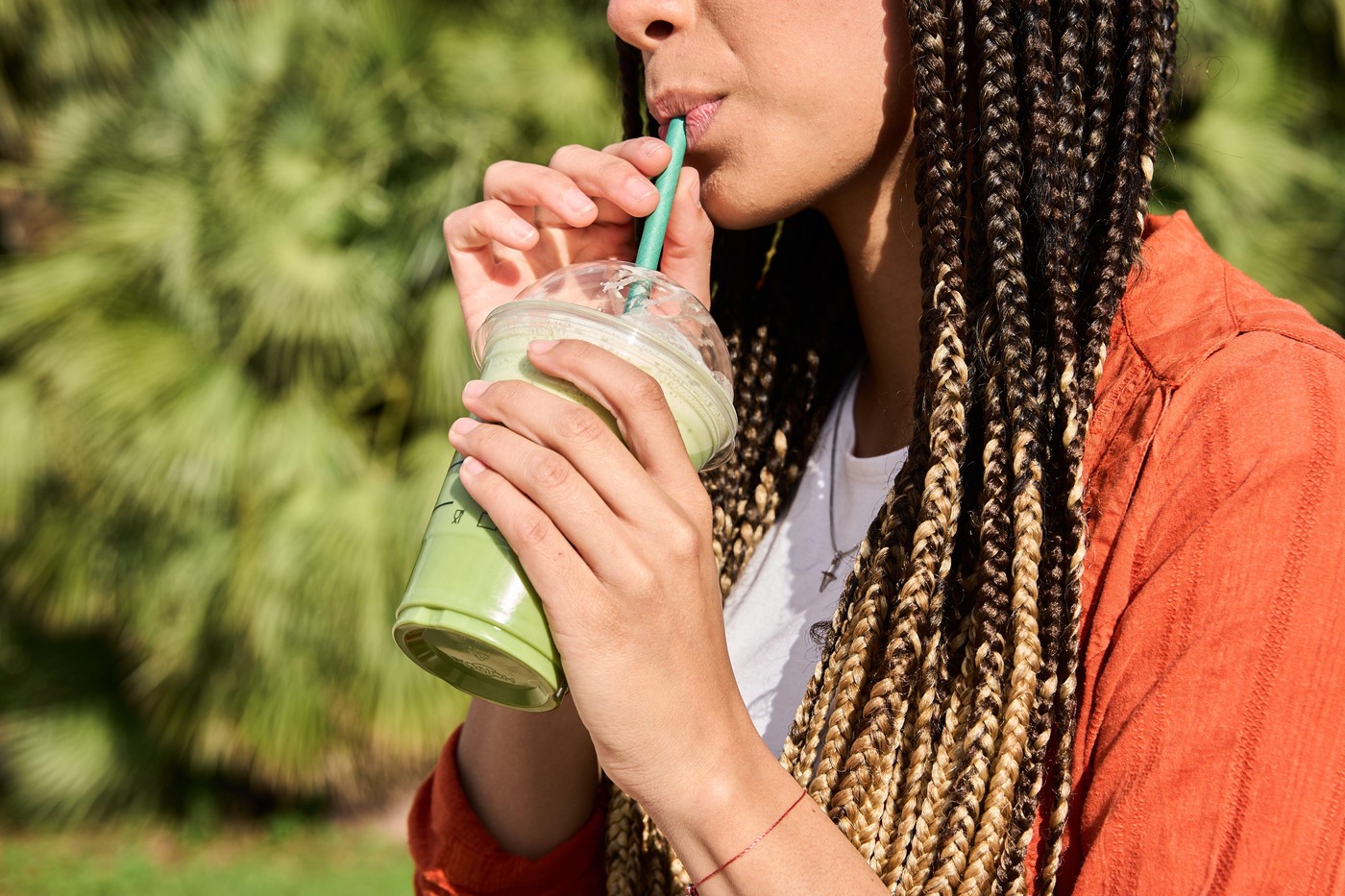 Young latina woman with long braids enjoys a refreshing green beverage through a straw on a sunny day outdoors