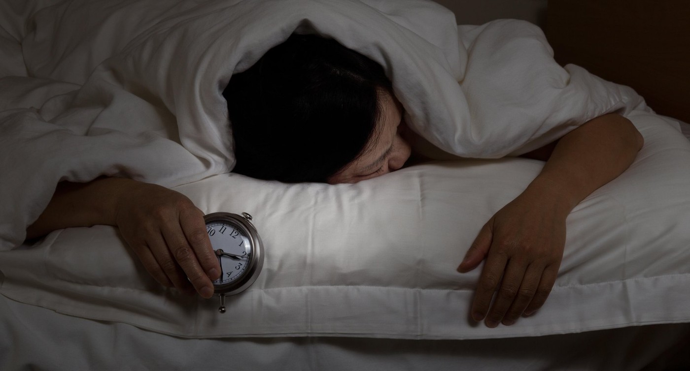 Woman with insomnia, under blanket looking down, holding alarm clock in hand. Select light and focus on woman and clock