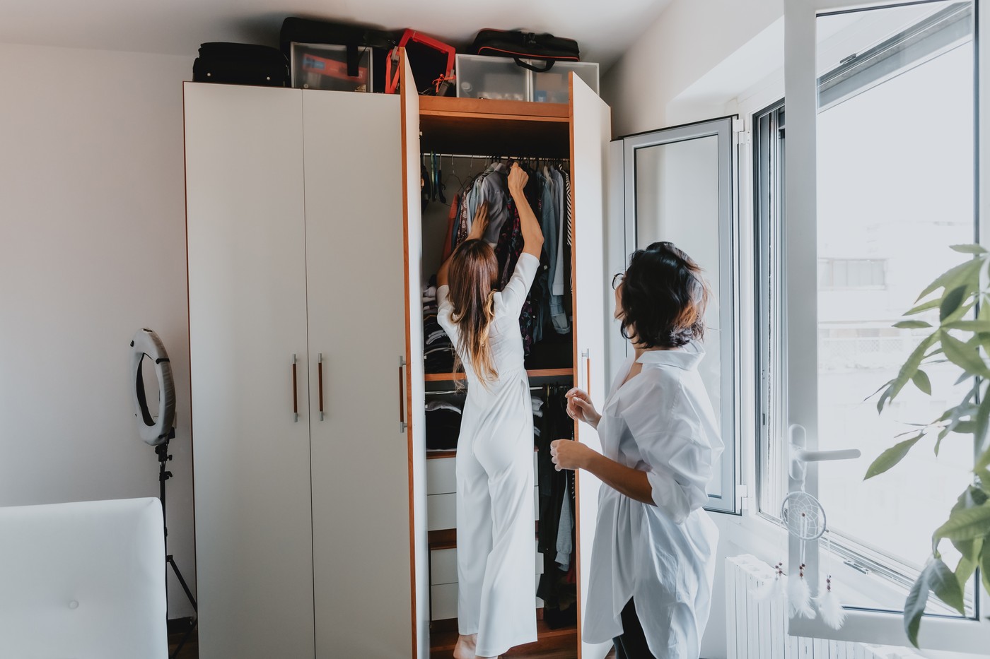 Two women with brown hair standing in an apartment, hanging clothes in wardrobe.