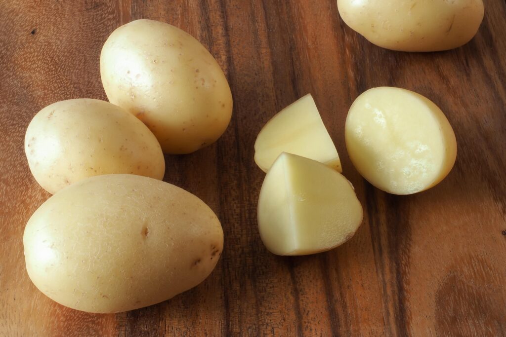 A small pale skinned potato cut into pieces, on a dark colored wooden cutting board, with whole potatoes surrounding.,Image: 288572964, License: Royalty-free, Restrictions: , Model Release: no, Credit line: Ralph Gillen / Alamy / Alamy / Profimedia