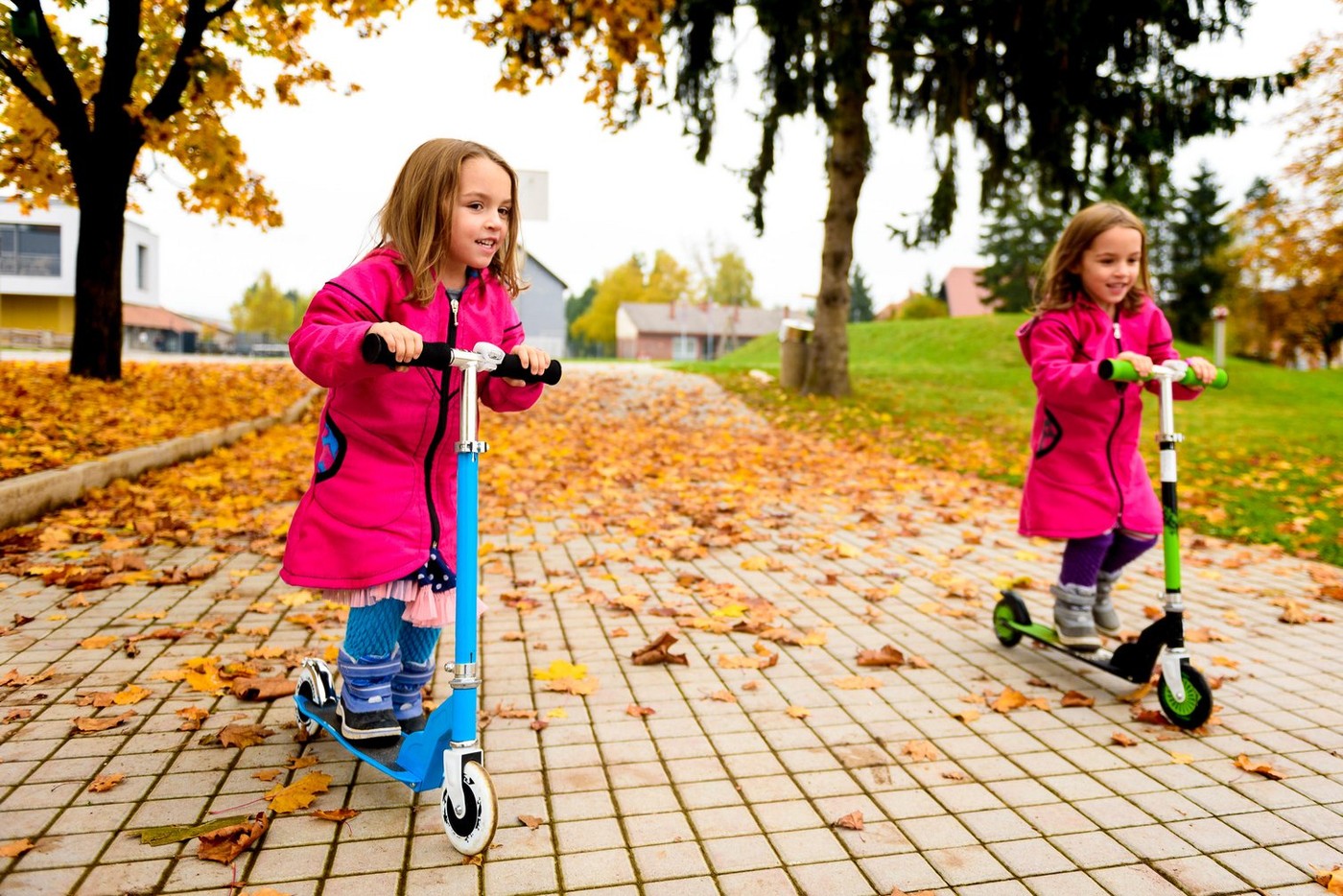 Twin girls in pink coat are riding scooter on maple leaves. Active children in sport pursuit is exercising in the nature in the autumn with cold tempe