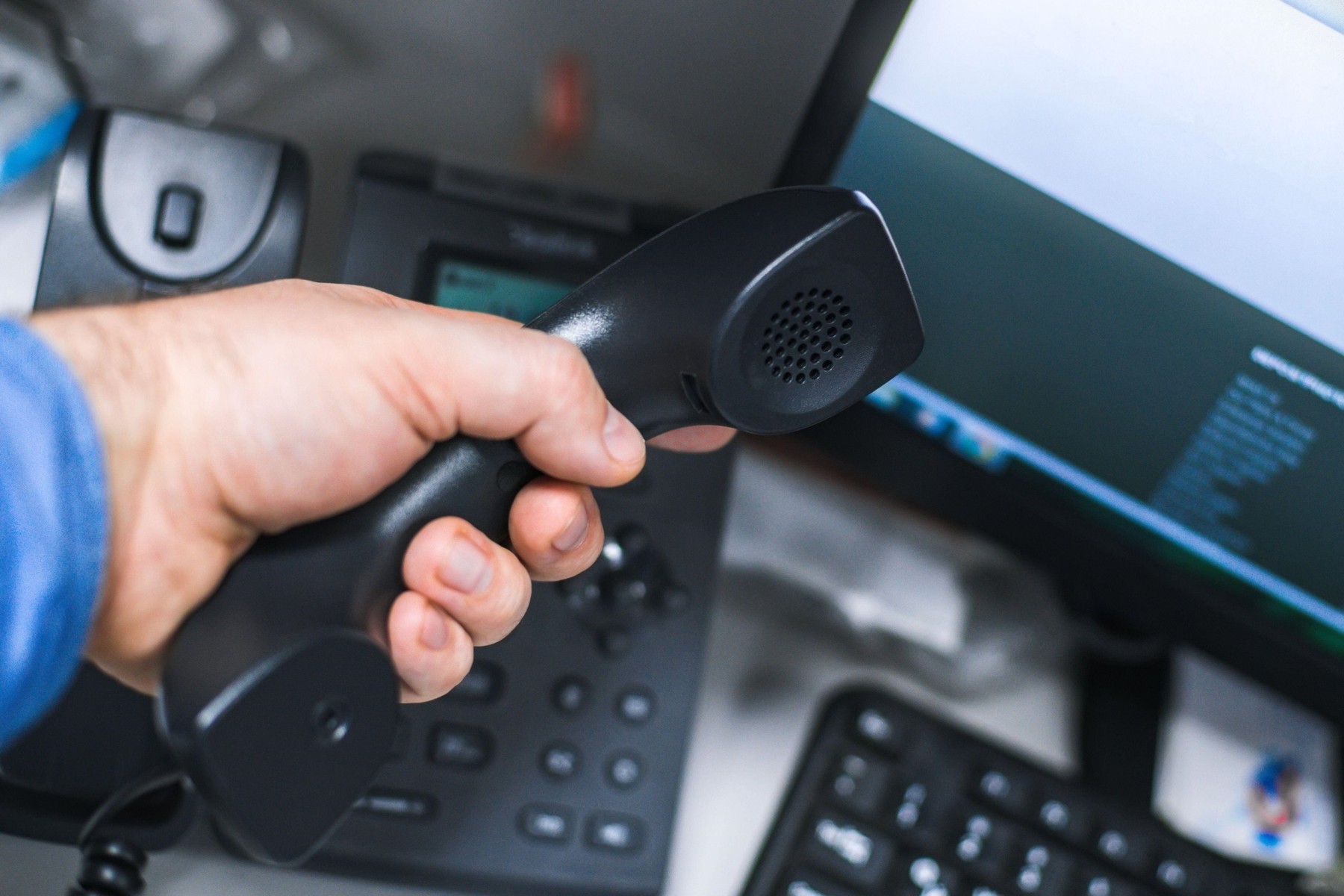 handset in hand and telephone with monitor and computer keyboard at shallow depth of field