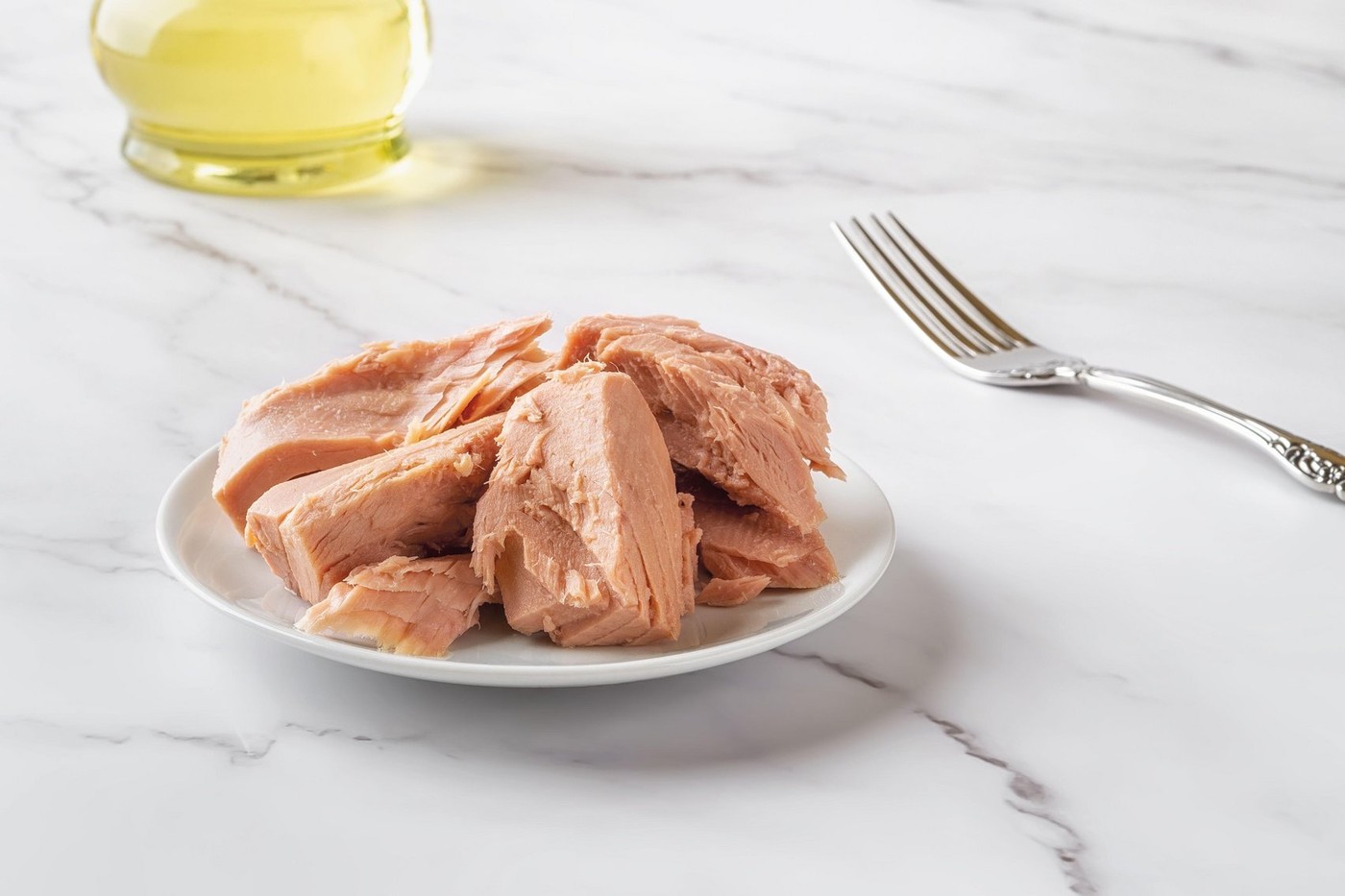 Canned tuna fillet pieces on a white saucer and fork over marble table close-up. Preserved tuna fish meat for low calorie diet eating. Tasty seafood.