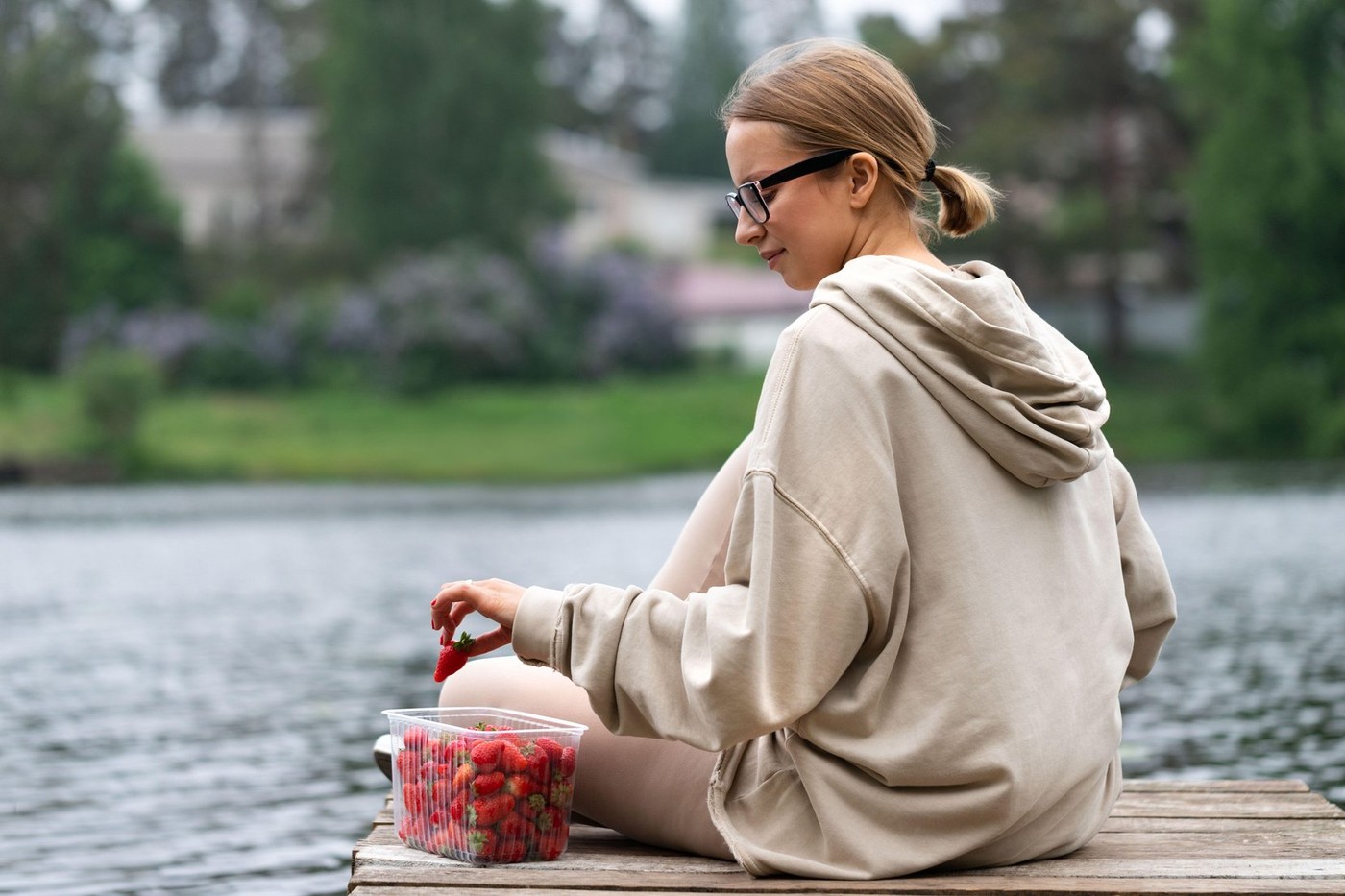 Young woman eating fresh berry fruit strawberries in plastic container, resting on a river wooden pier, back view. Summer time. The joy of country lif