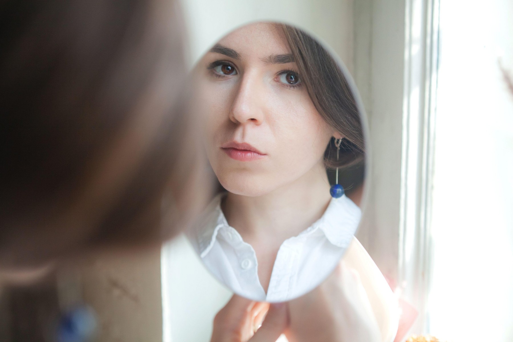 young beautiful girl with brown eyes and a blue earring looks in the mirror. Portrait reflection in the mirror of a sad woman