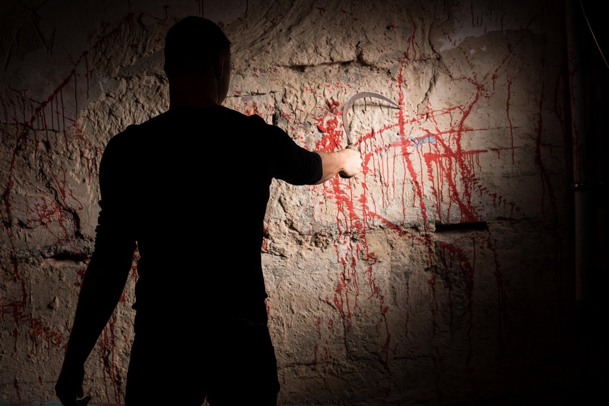 Portrait of shadowy male figure holding blade and scratching blood stained wall for concept about murder and scary Halloween hol