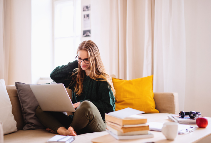 PR tekst  A young happy college female student sitting on sofa at home, using laptop when studying.