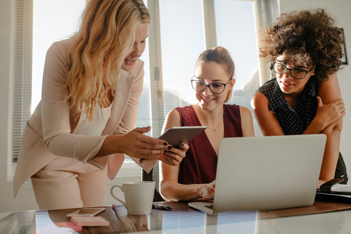 PR tekst  Three beautiful creative business woman working with laptop and watching something on tablet in office. Group of businesswoman working over a new project at startup office.
