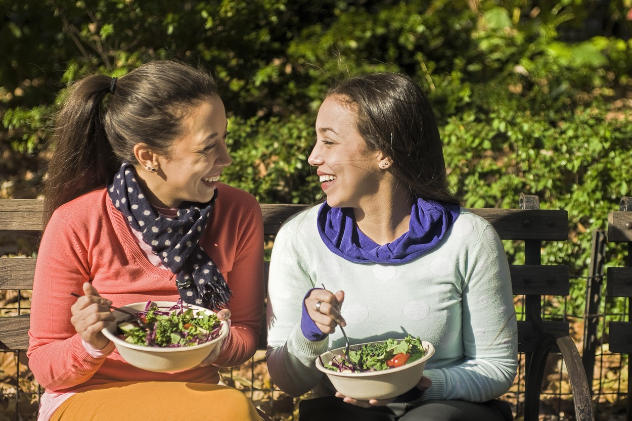 Young adult female twins sitting on park bench eating lunch