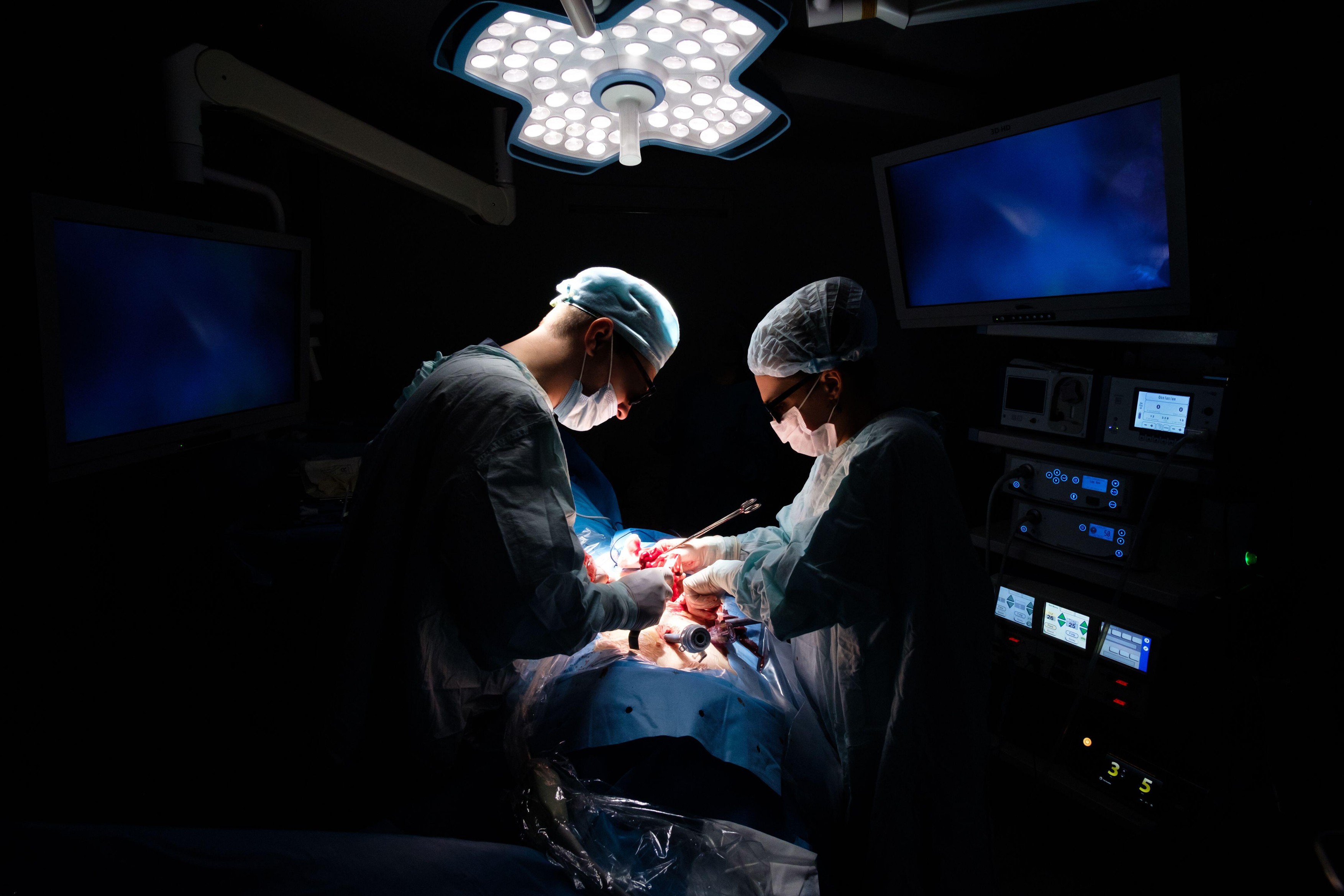 A team of surgeons wearing 3D glasses operate on a patient in the bright light of operating lamps. Laparoscopic minimally invasive surgery. Monitors of modern medical equipment glow in the dark.