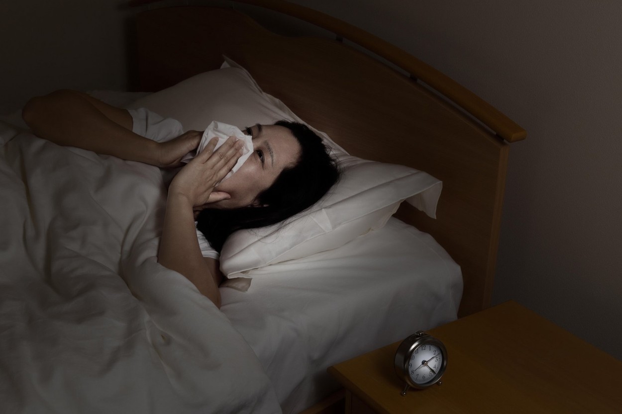 Woman wiping nose with tissue while trying to sleep. Select light and focus on woman and clock with darker background. Sickness