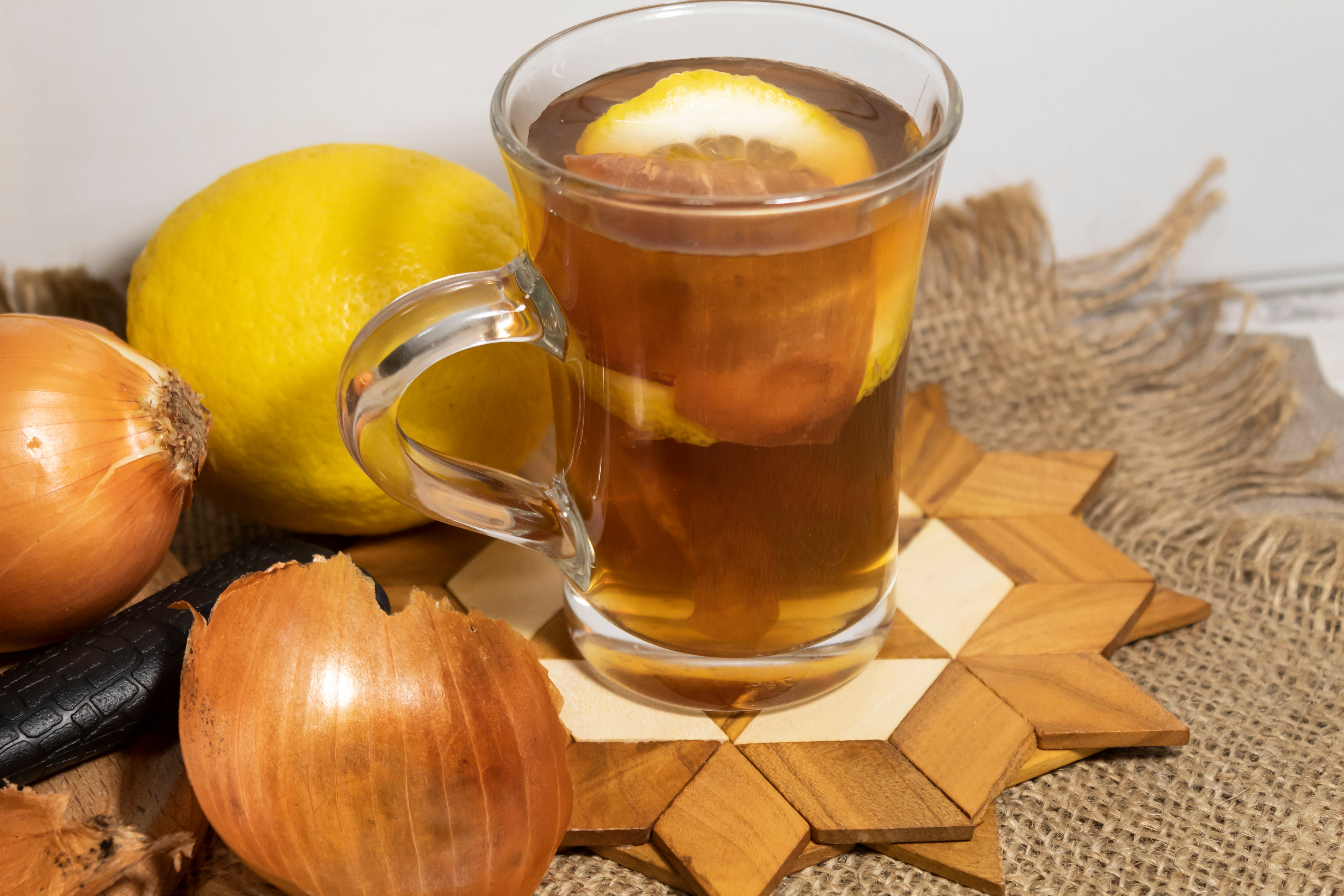 On a jute napkin, on a stand, there is a cup with brewed onion peels and lemon. Onions and lemon lie nearby.