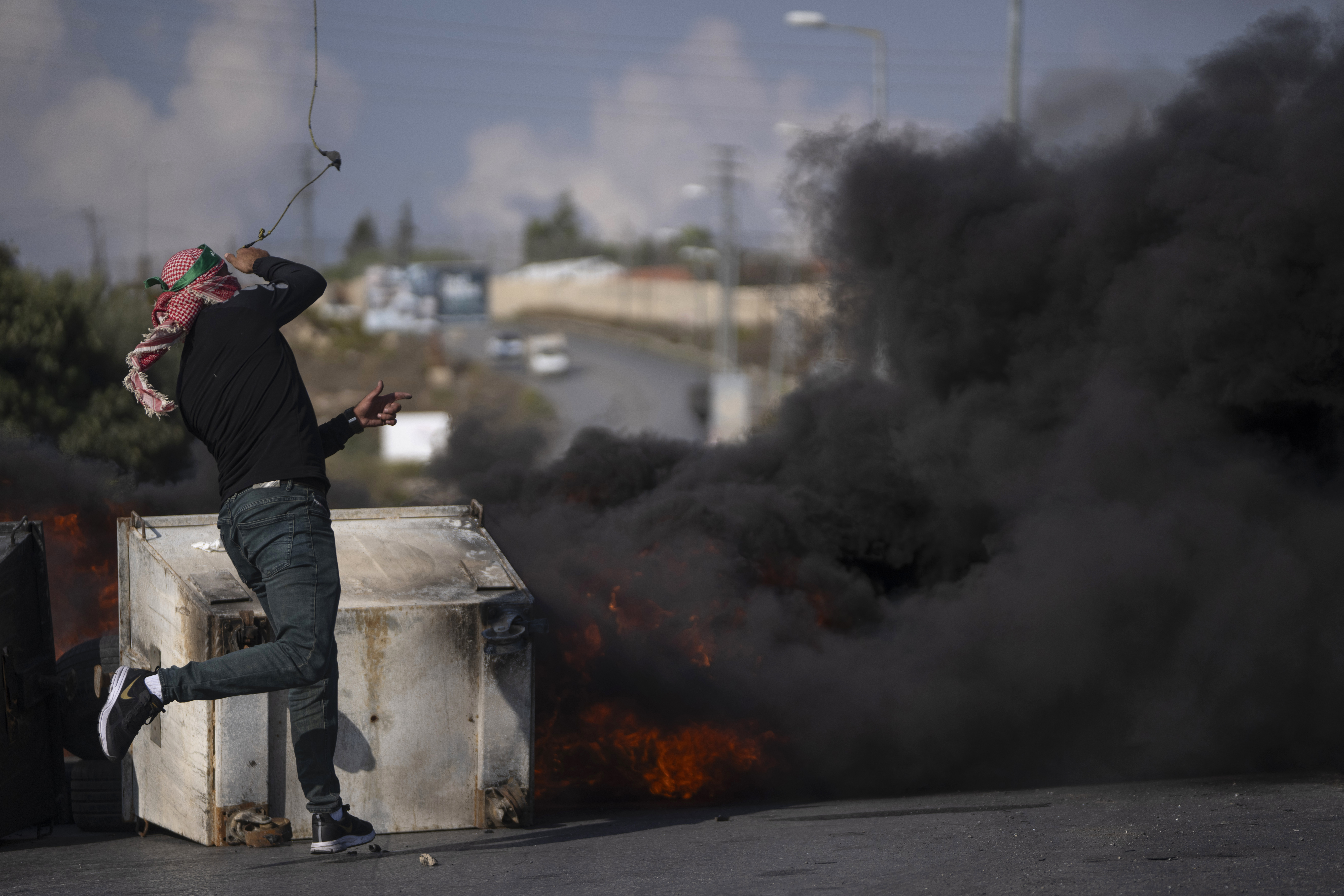A Palestinian protester uses a slingshot behind burning tires during clashes with Israeli border police following a protest against Israeli airstrikes on Gaza, in the West Bank city of Ramallah, Wednesday, Oct. 18, 2023. (AP Photo/Nasser Nasser)