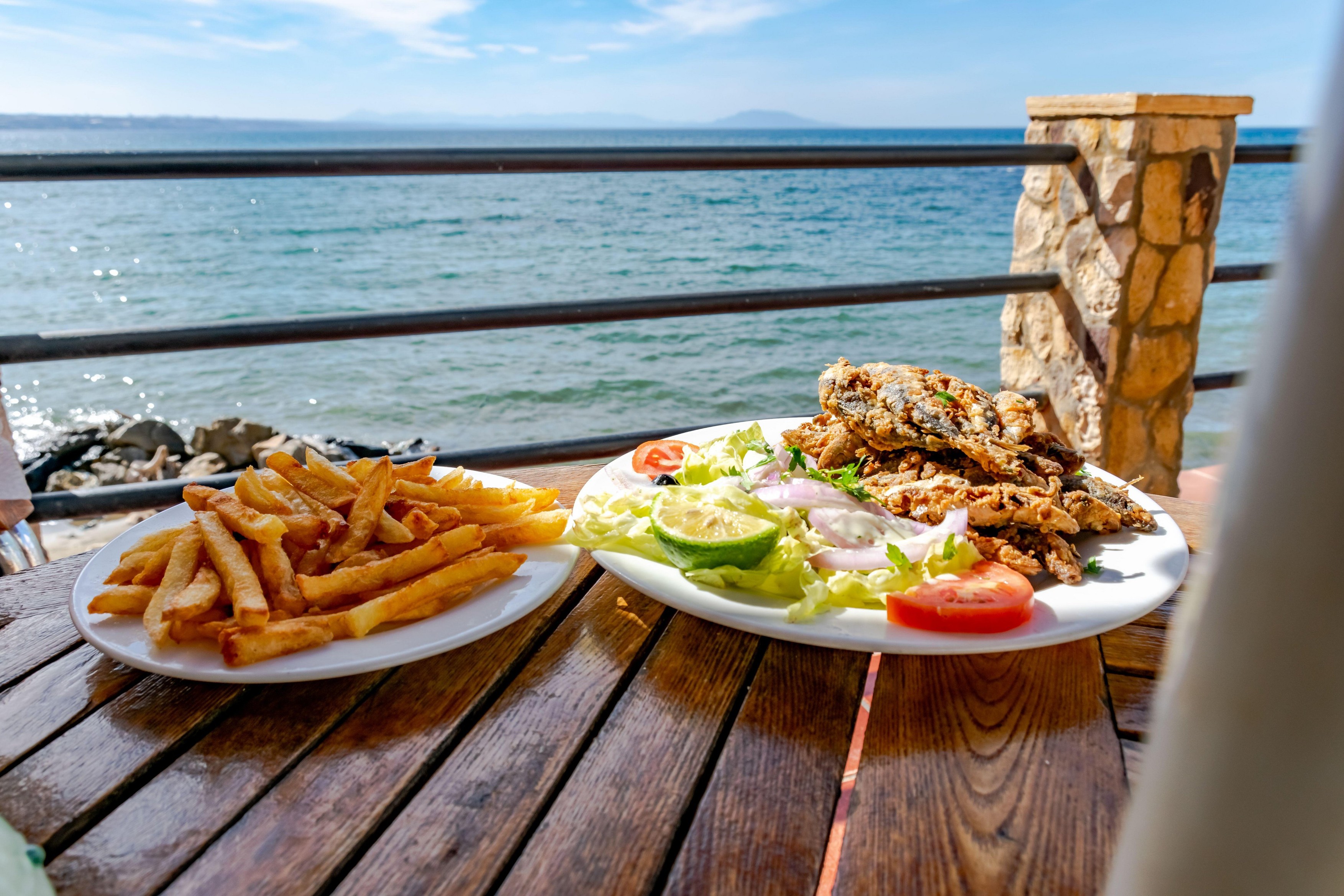 A plate of French fries and grilled sardines with salad, tomato and lemon. Restaurant in edge of the Mediterranean sea with a view from the balcony.