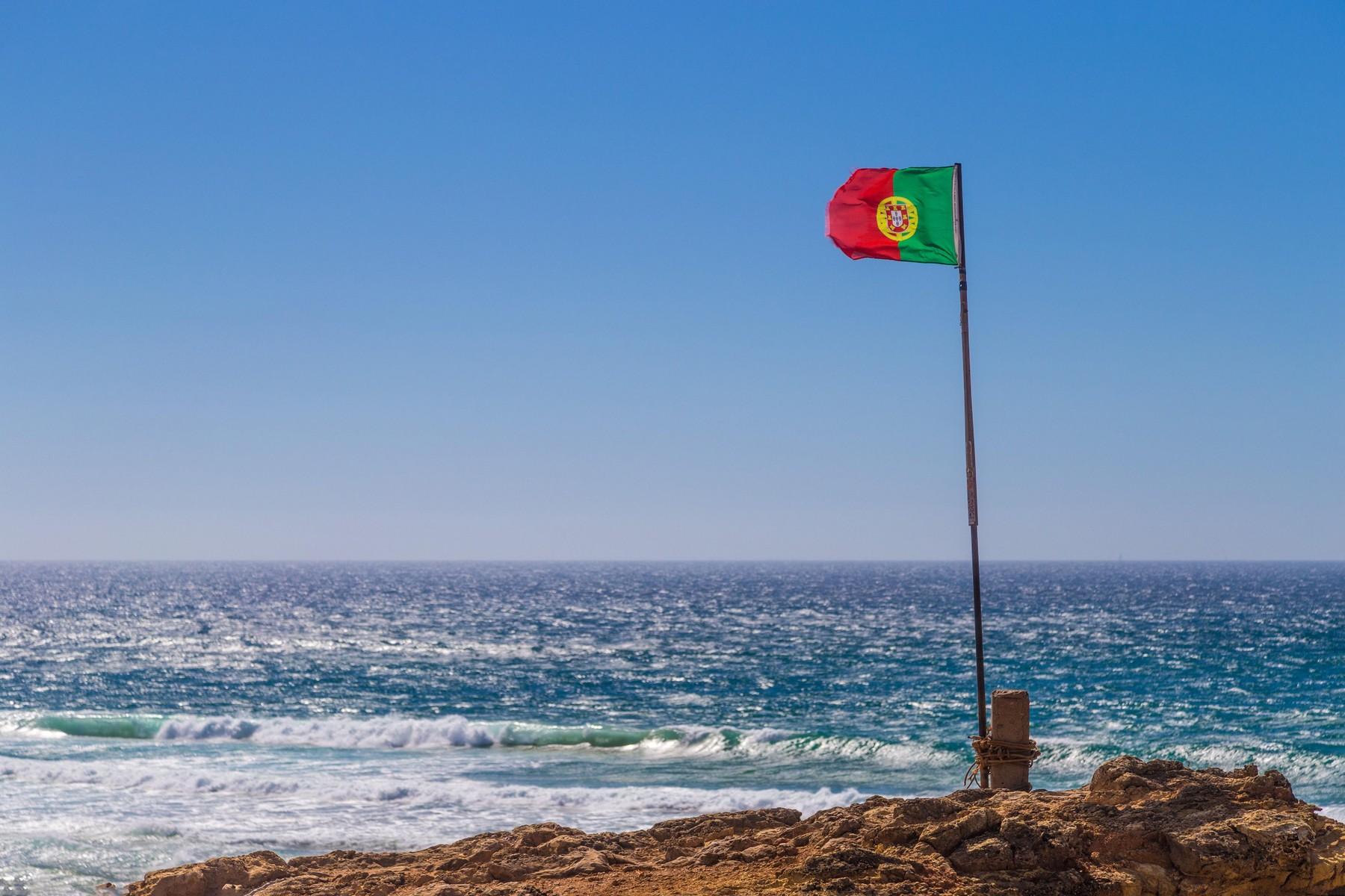Portuguese flag waving over blue sky and ocean, Guincho Beach, Portugal