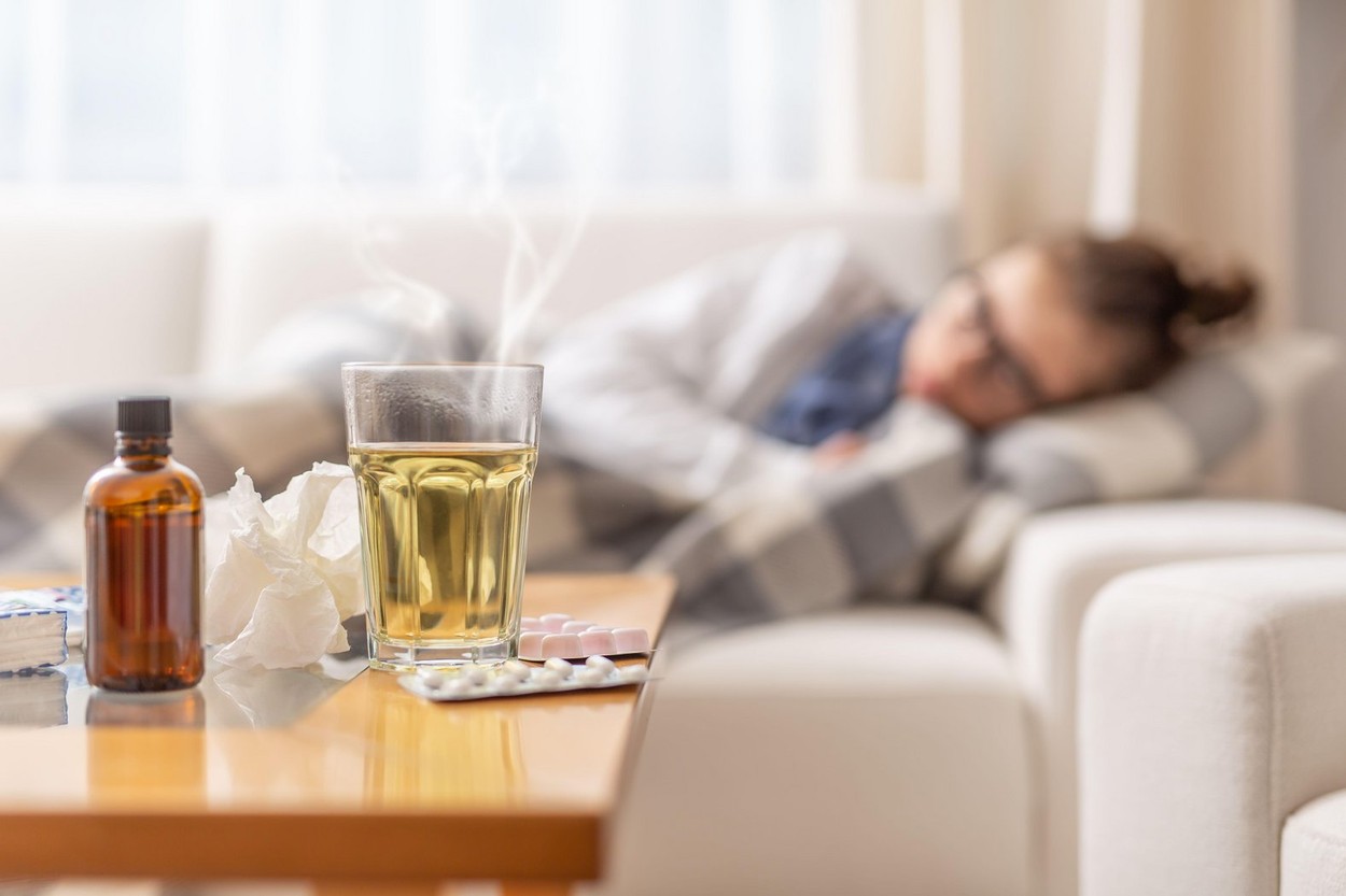 Young ill woman lying under blanket on a bed with seasonal illnes drinking hot tea with honey and lemon. She takes pills and cough sirup to push the d