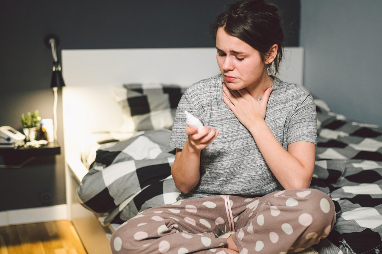 The theme is seasonal cold, runny nose flu virus infection. Young caucasian woman at home bedroom bed uses spray drops of medicine in her nose for sno