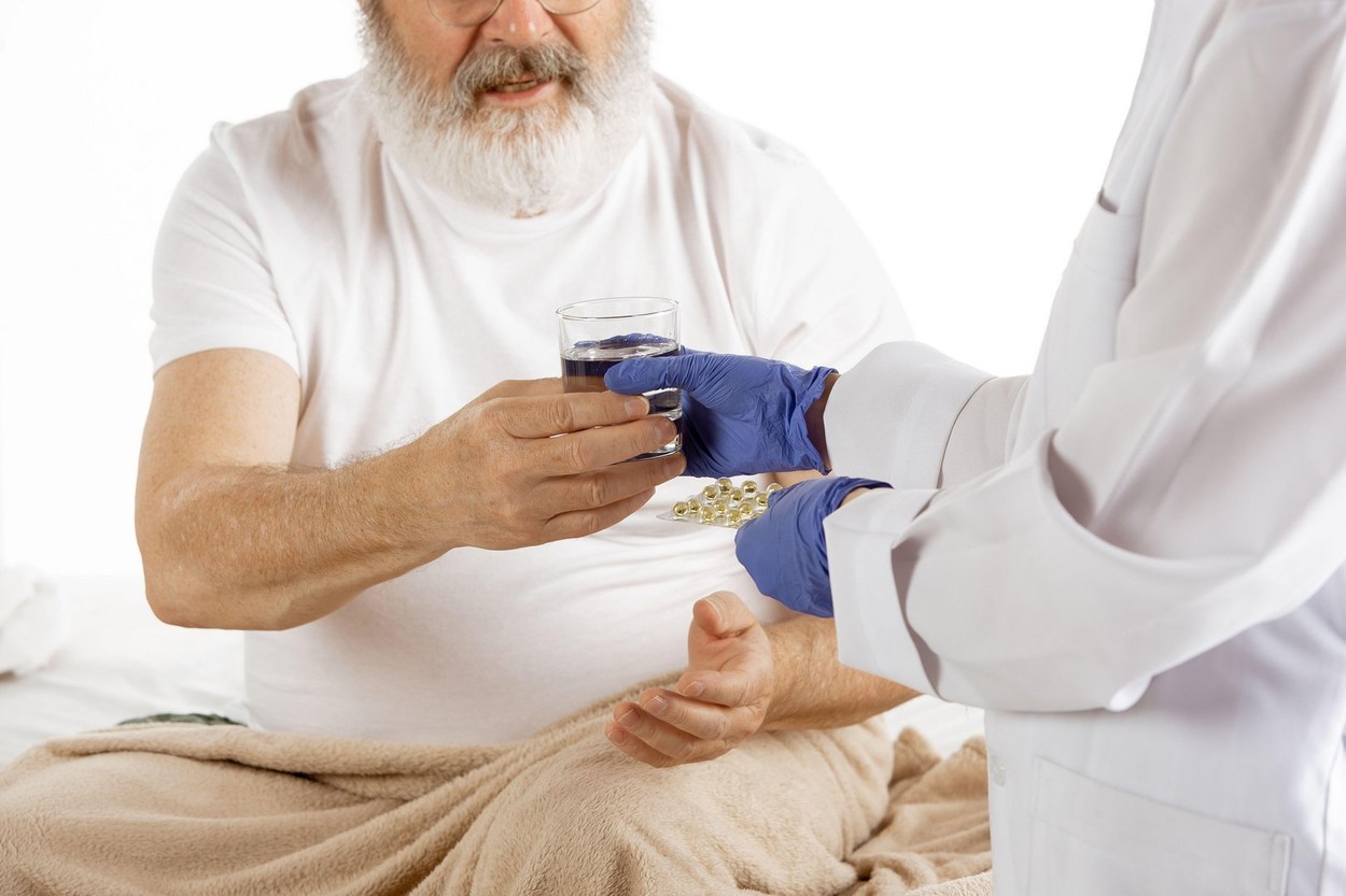 Elderly old man recovering in a hospital bed isolated on white background. Getting care and treatment. Concept of healthcare and medicine. Close up nurse giving pills and water. Copyspace.
