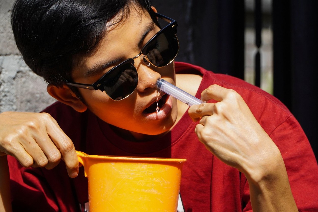 A woman with short hair wearing sunglasses and a red shirt, is using a syringe to inject NaCL fluid into her nose to clean her nose of germs and disea
