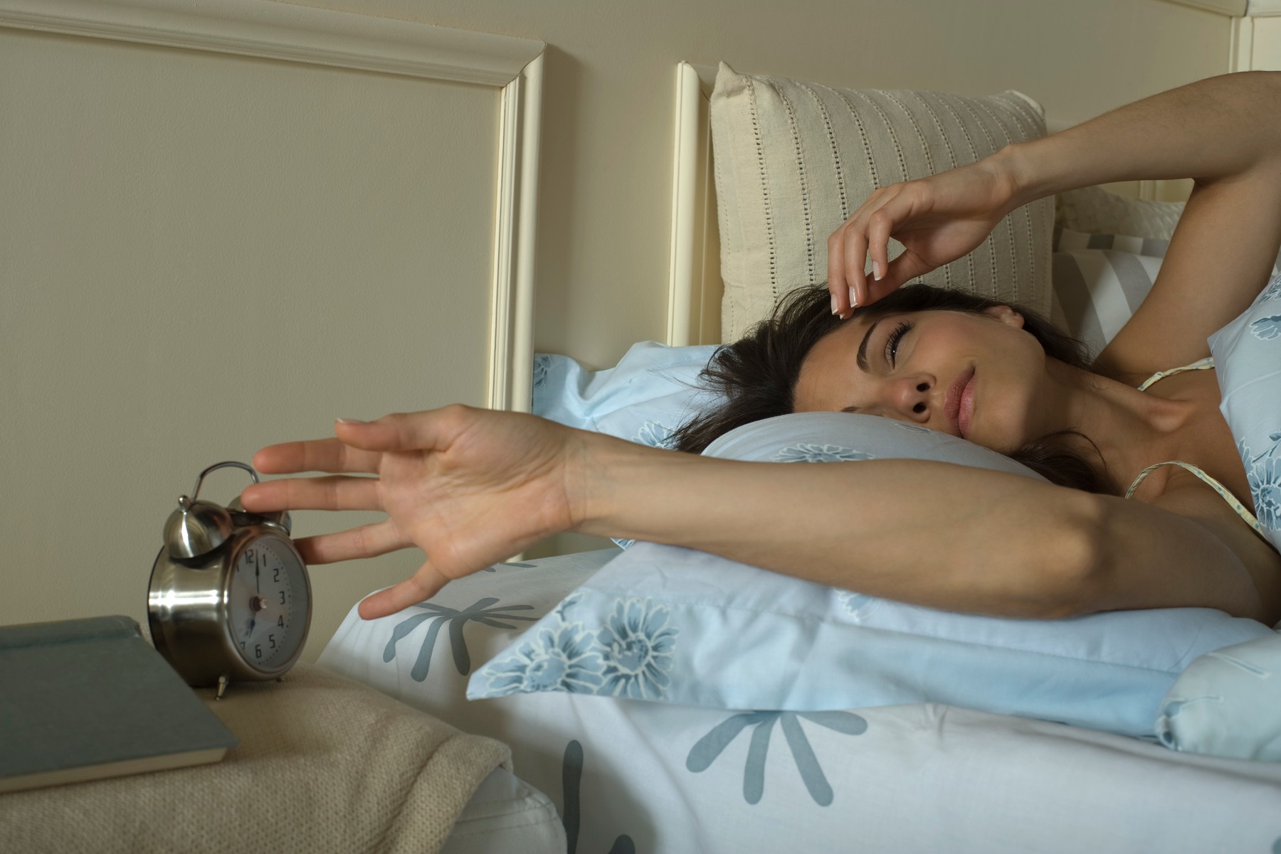 Young woman lying in bed, silencing alarm clock