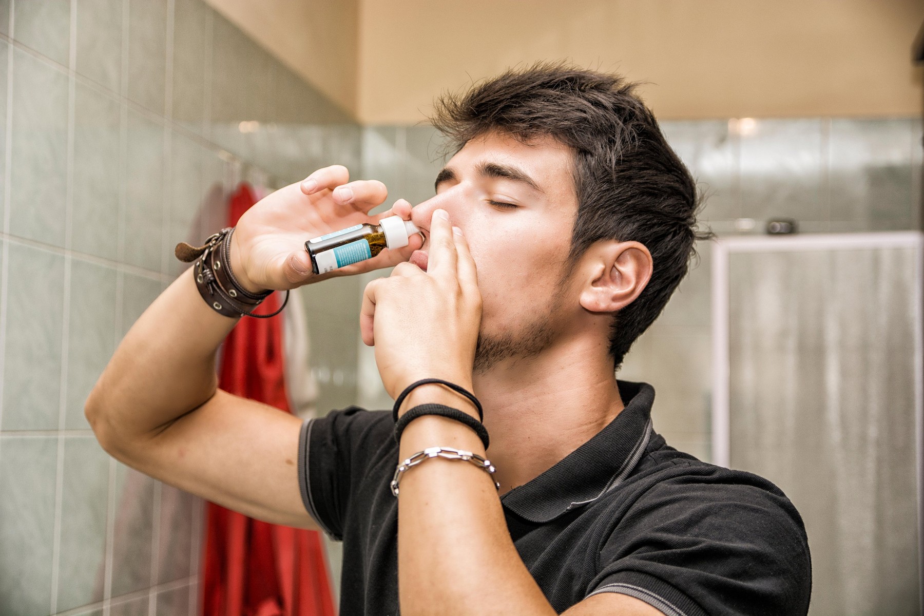 Head and Shoulders Close Up of Young Attractive Man with Dark Hair Sniffing Nose Spray with Eyes Closed in Home Bathroom