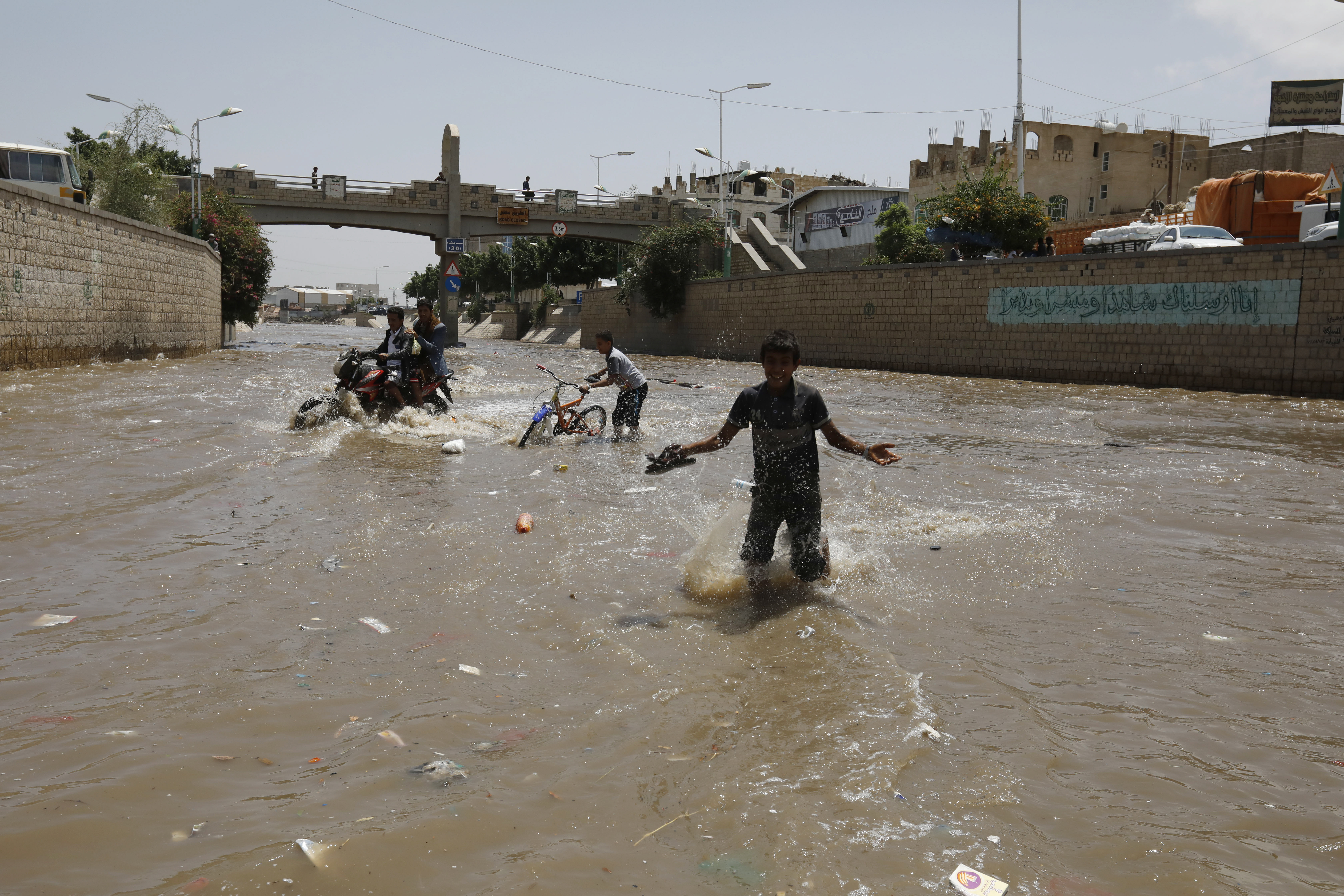 epa08559169 People wade and attempt to traverse a flooded street with a motorcycle and a bicycle following heavy rainfall in Sana'a, Yemen, 21 July 2020. Strong monsoon rains have affected most parts of the war-torn nation.  EPA-EFE/YAHYA ARHAB