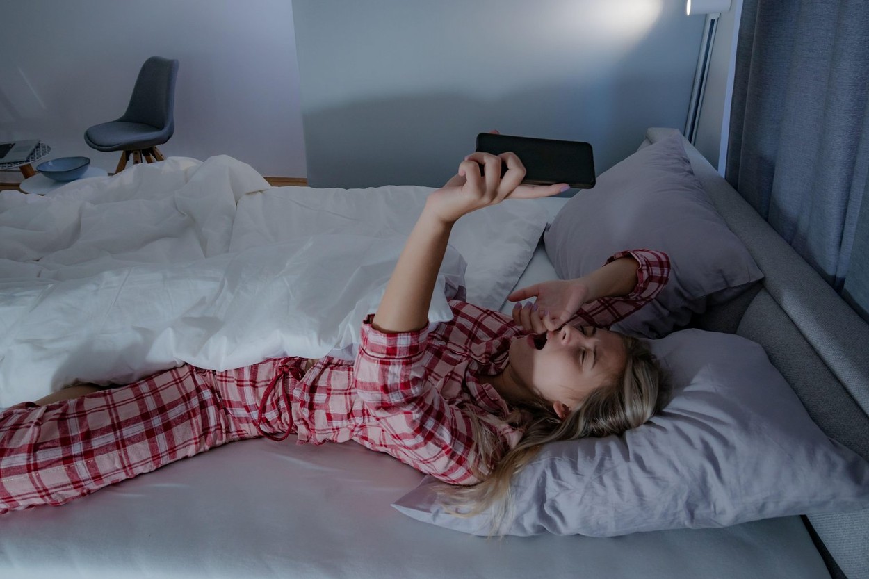 Beautiful young woman in pajamas stretching in bed, turning off the alarm clock on a mobile phone, having a hard time to wake up.