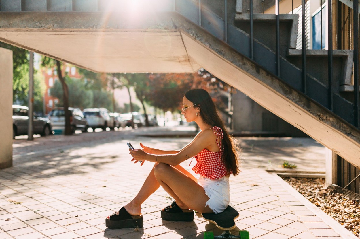 Serious and thoughtful woman sitting on a skateboard with a cellphone in her hands