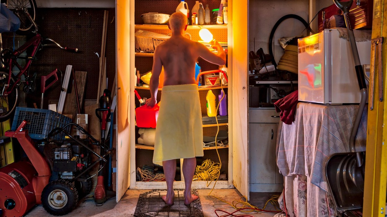 A bachelor man wearing only a towel, in the garage searching for something in a closet.