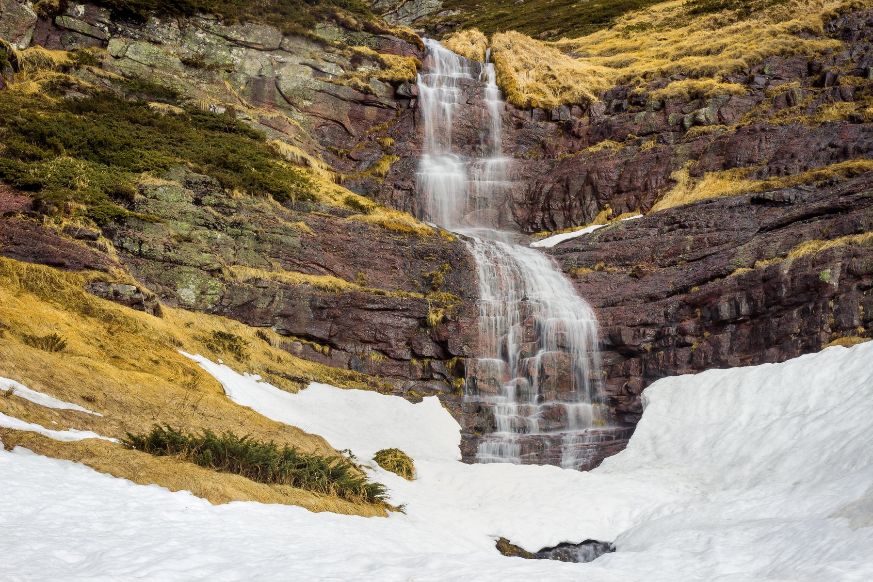 Amazing, unique Kopren waterfall streaming down the red rocks of a vertical cliff covered by yellow, dry grass