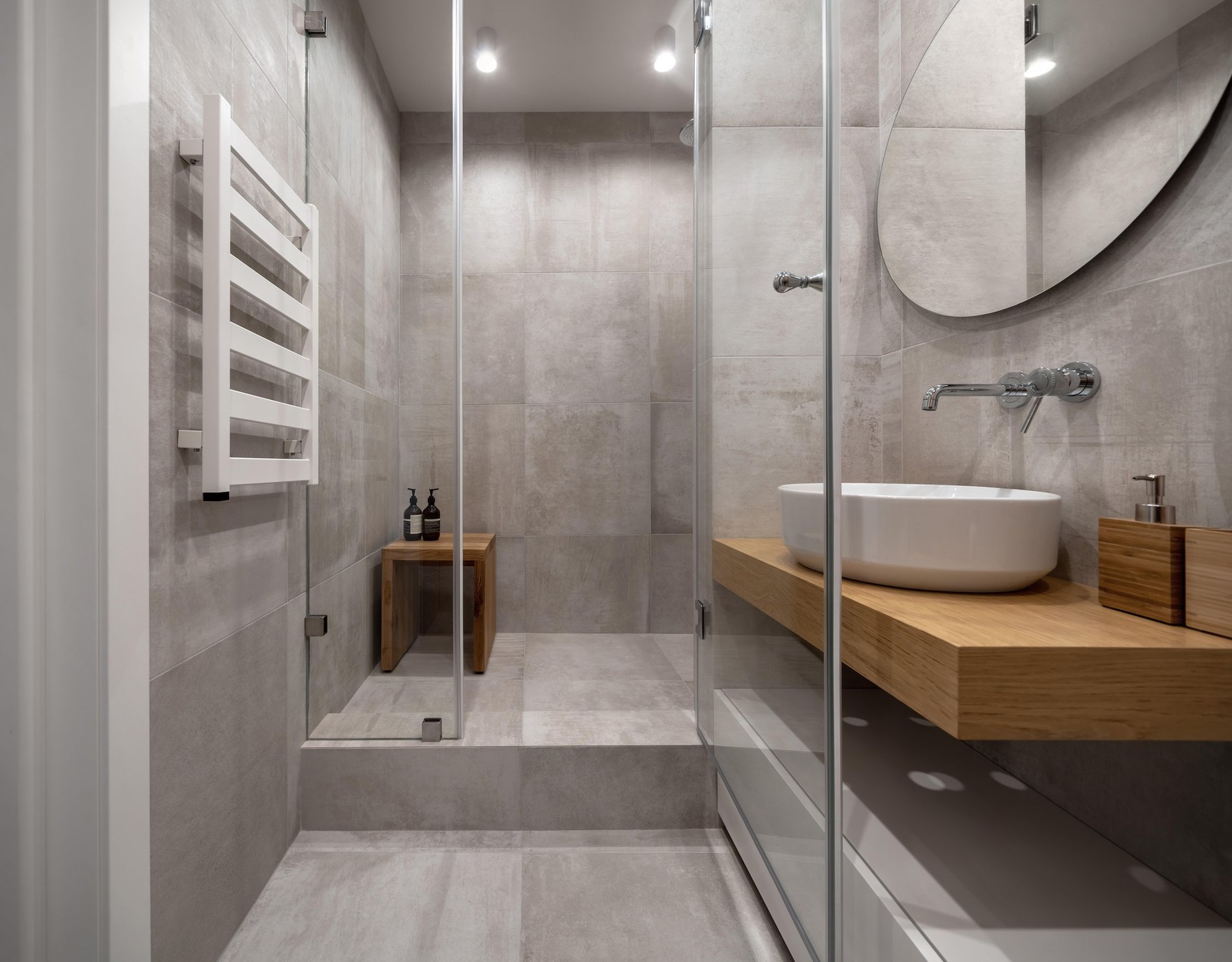 Bathroom in a modern style with textured beige tiled walls and luminous lamps. There is a glass shower cabin, wooden rack with a white sink and soap d