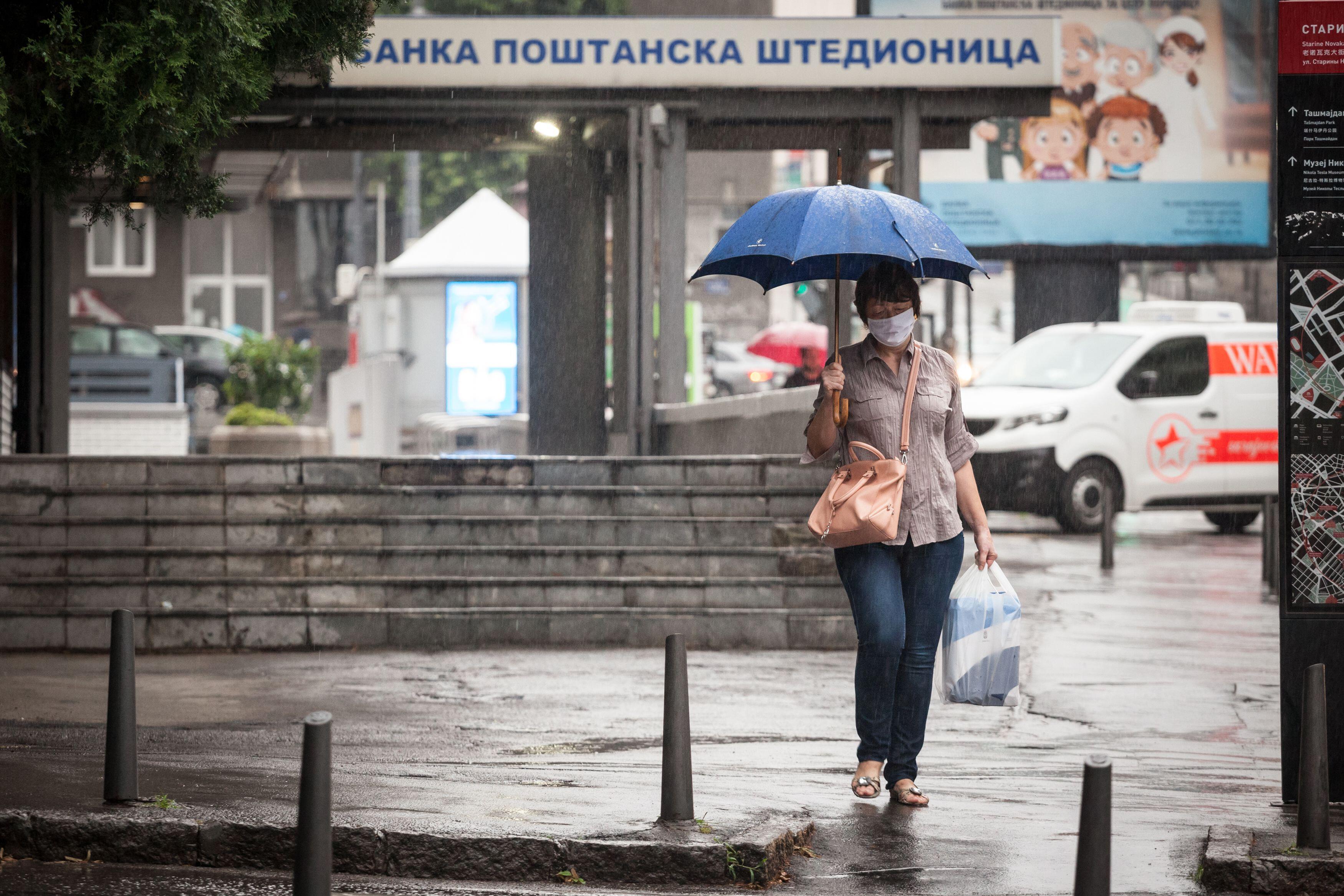 BELGRADE, SERBIA - JULY 17, 2020: Old senior woman wearing a respiratory face mask walking with her umbrella under heavy rain in a street of Belgrade,