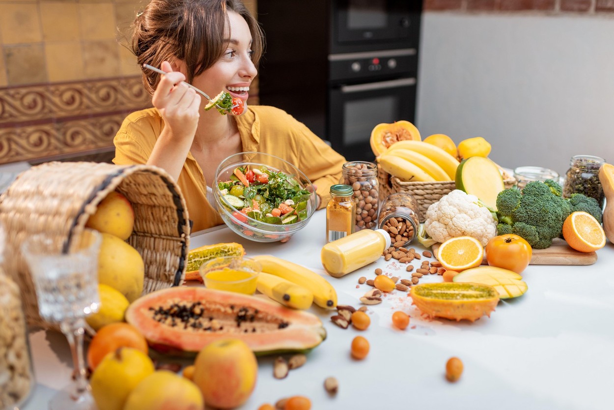 Portrait of a young cheerful woman eating salad at the table full of healthy raw vegetables and fruits on the kitchen at home. Concept of vegetarianism, healthy eating and wellness