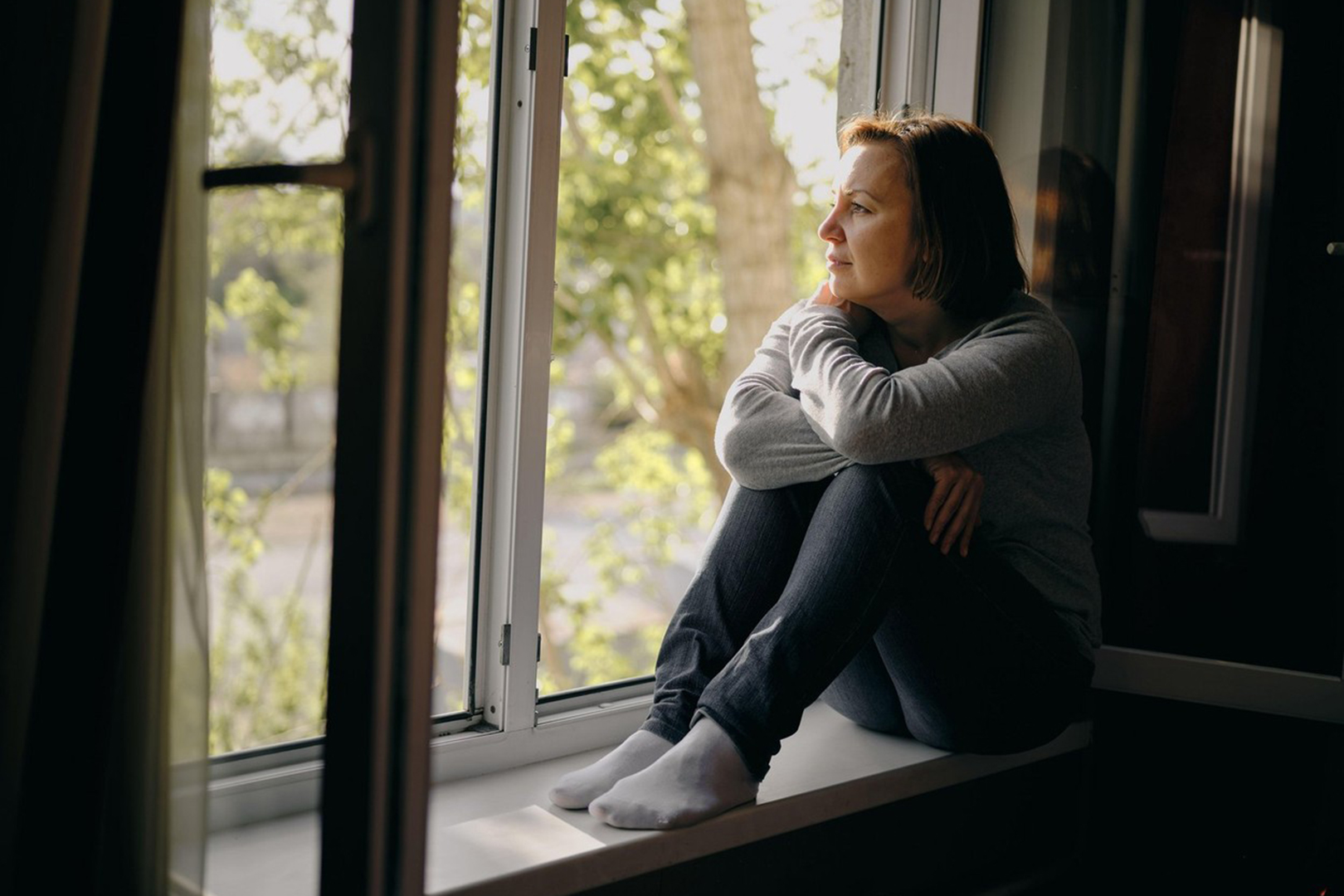 A beautiful young woman is sad sitting on a window in her house. Quarantine and self-isolation due to the coronavirus pandemic.
