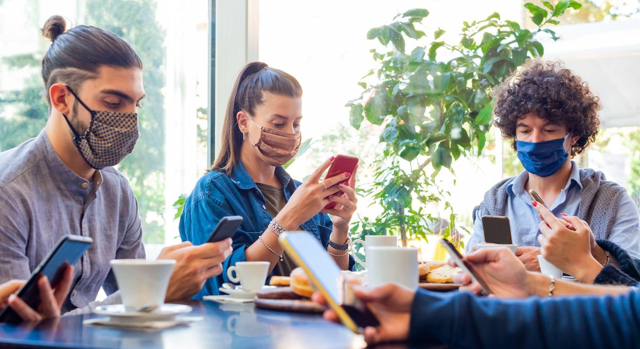 happy group of friends smiling at breakfast into a cafe whilst interacting with technology. mates drinking coffee and eating while using smartphones.