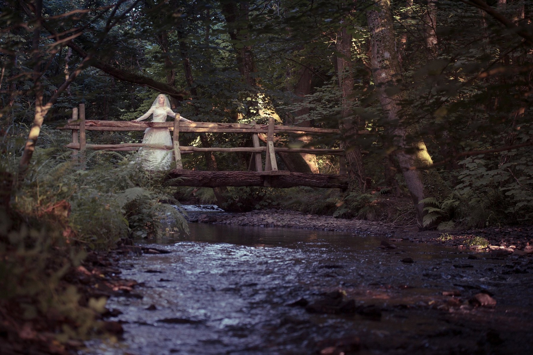 A young blonde woman girl wearing an old-fashioned white wedding dress frock standing on a rustic wooden bridge in a woodland on a summer evening,, UK