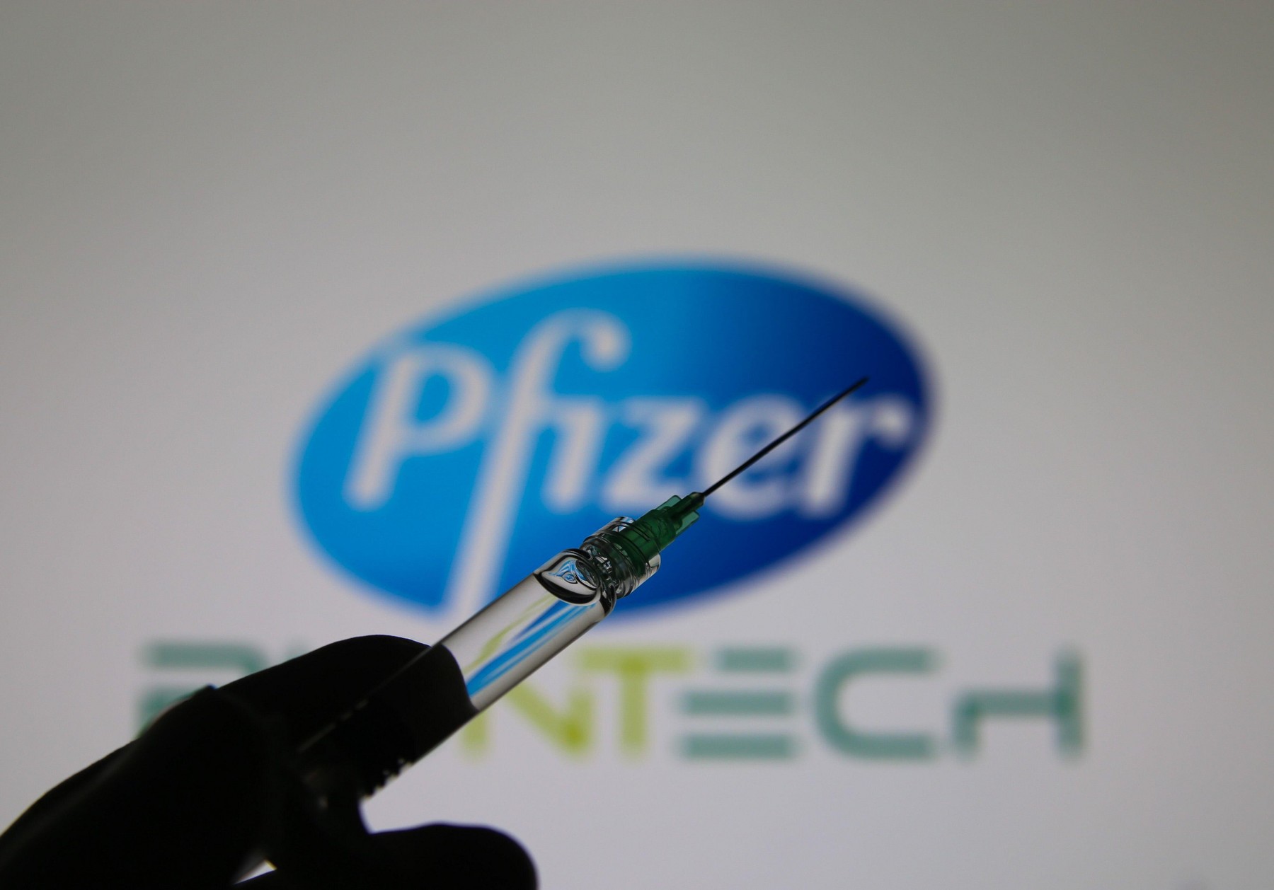 Viersen, Germany - November 9. 2020: Close up of syringe hold by hand with injection needle and serum, blurred Pfizer and Biontech logo lettering back