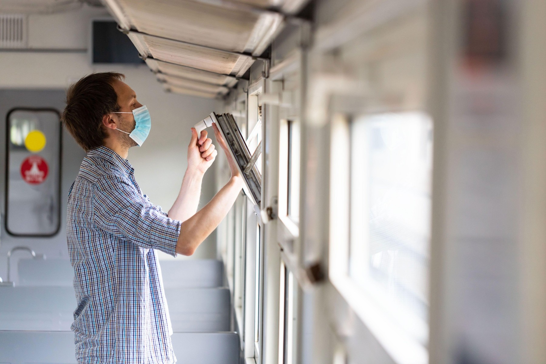 Man in checked shirt opens a window on a train to breathe fresh air and ventilation, wearing medical facial mask during new normal change after corona