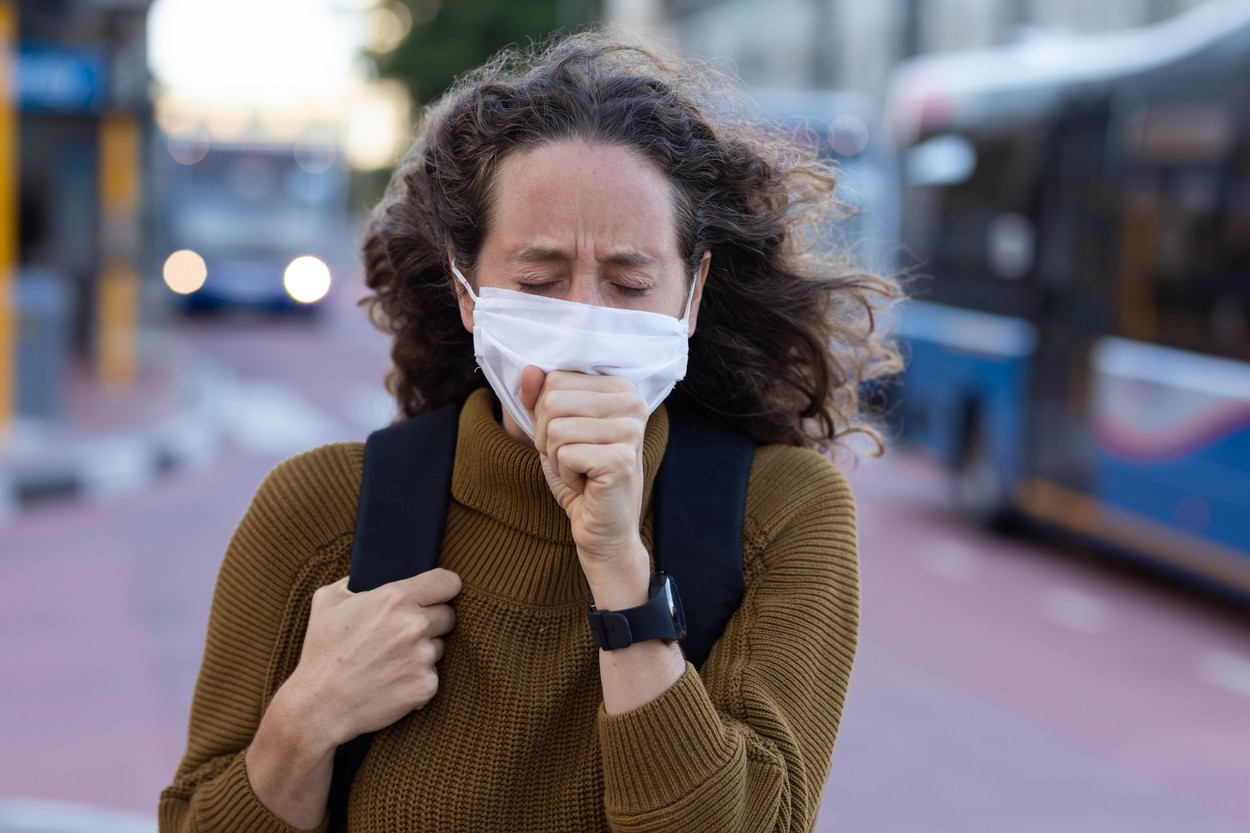 Caucasian woman out and about in the city streets during the day, wearing a face mask against covid19 coronavirus covering her face while coughing